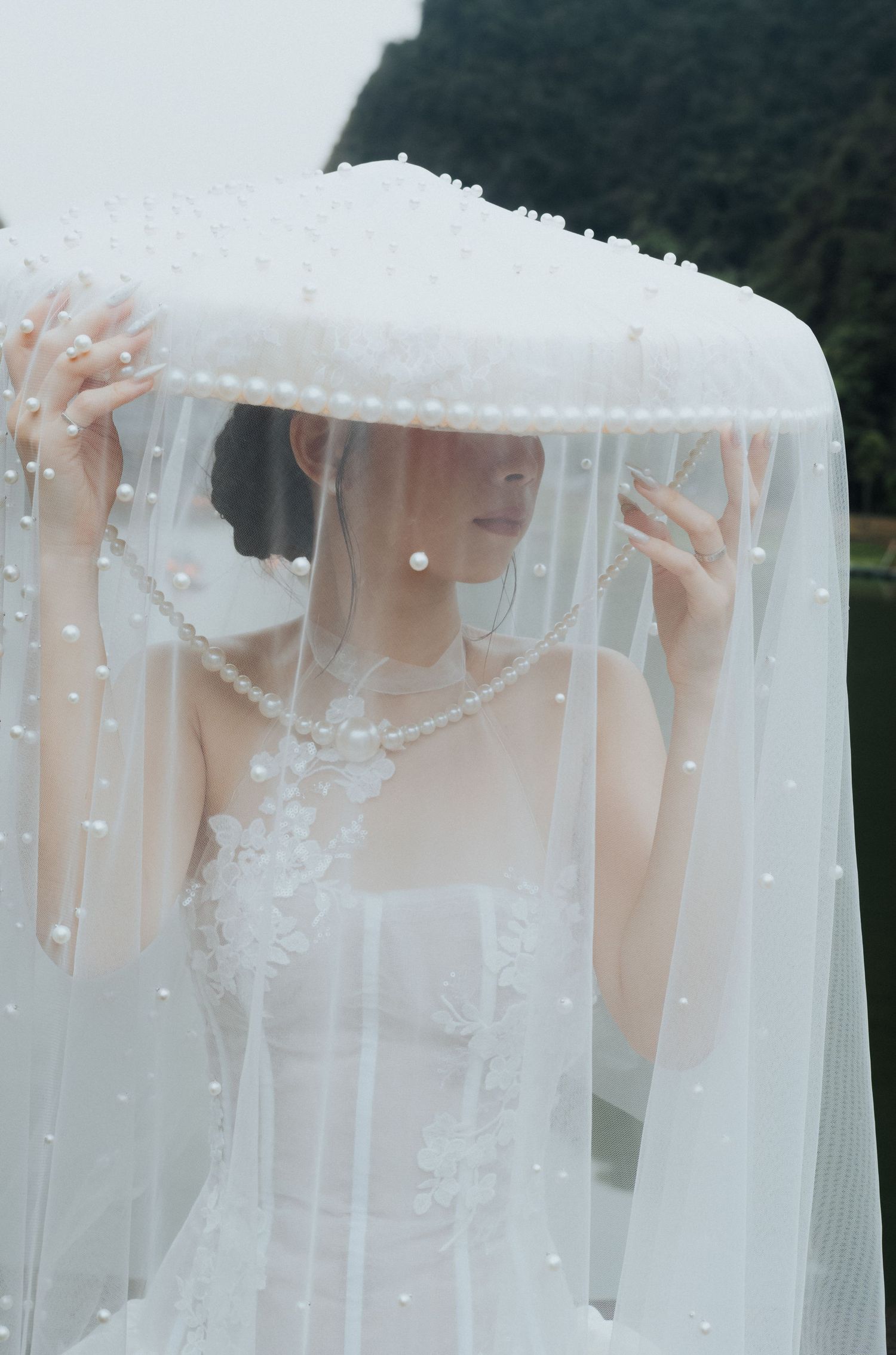 Bride in white lace gown holds pearl-trimmed veil overhead, wearing pearl necklace and earrings for elegant bridal portrait.