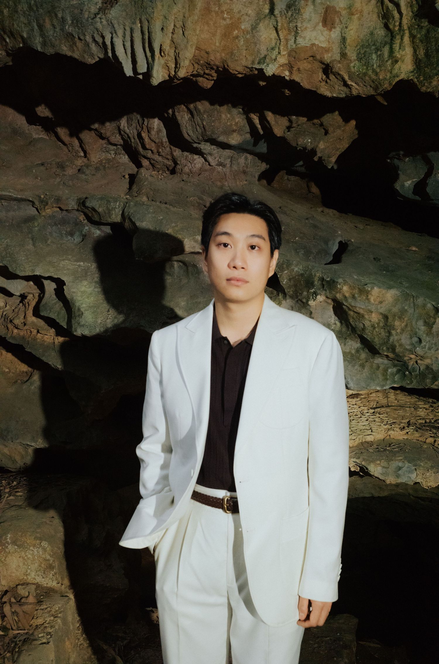 Young Asian man in white suit and black shirt poses confidently inside a dramatic rocky cave interior.