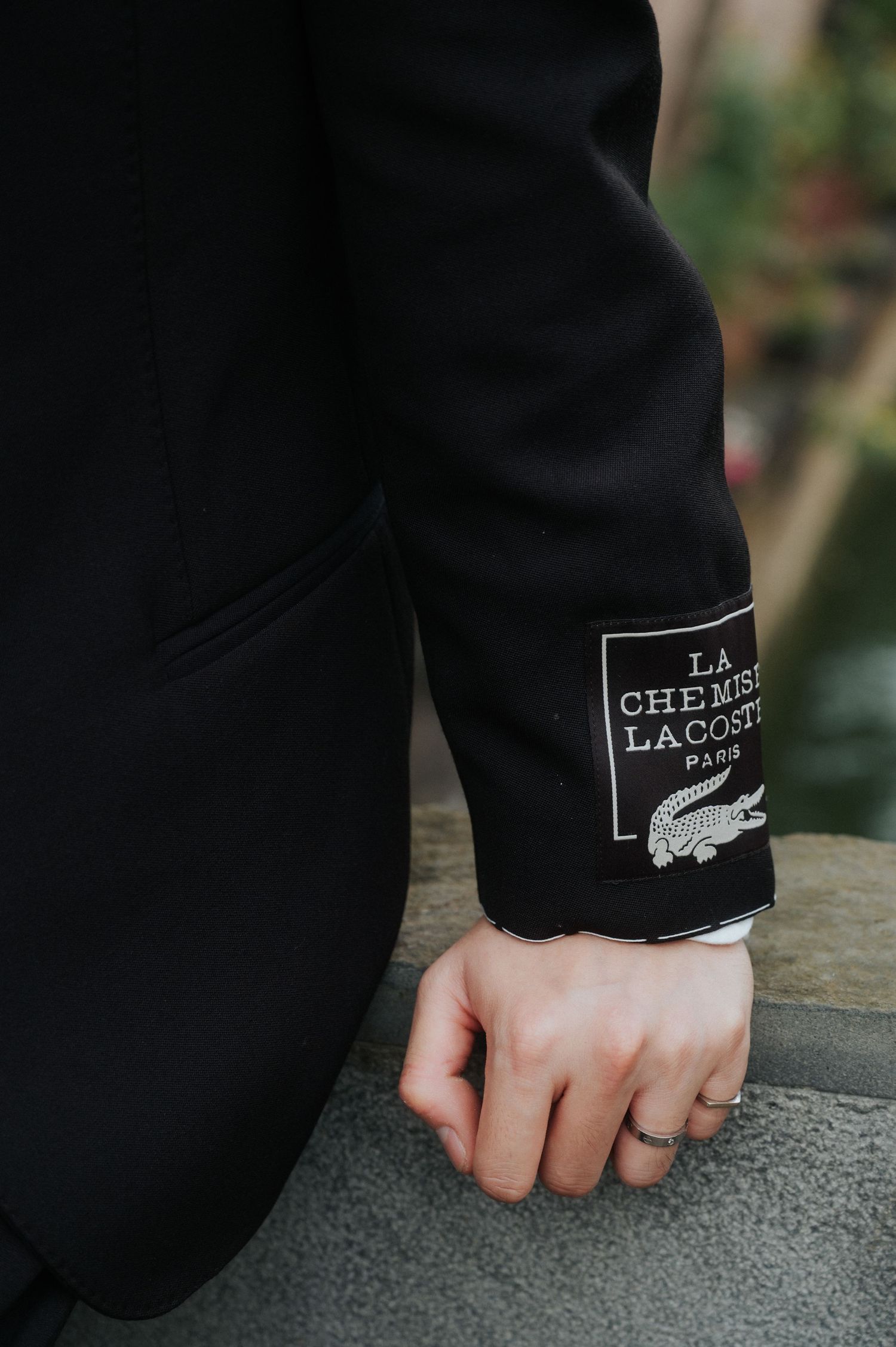 Close-up of a black Lacoste La Chemise Paris jacket sleeve with logo patch, worn by a person outdoors.