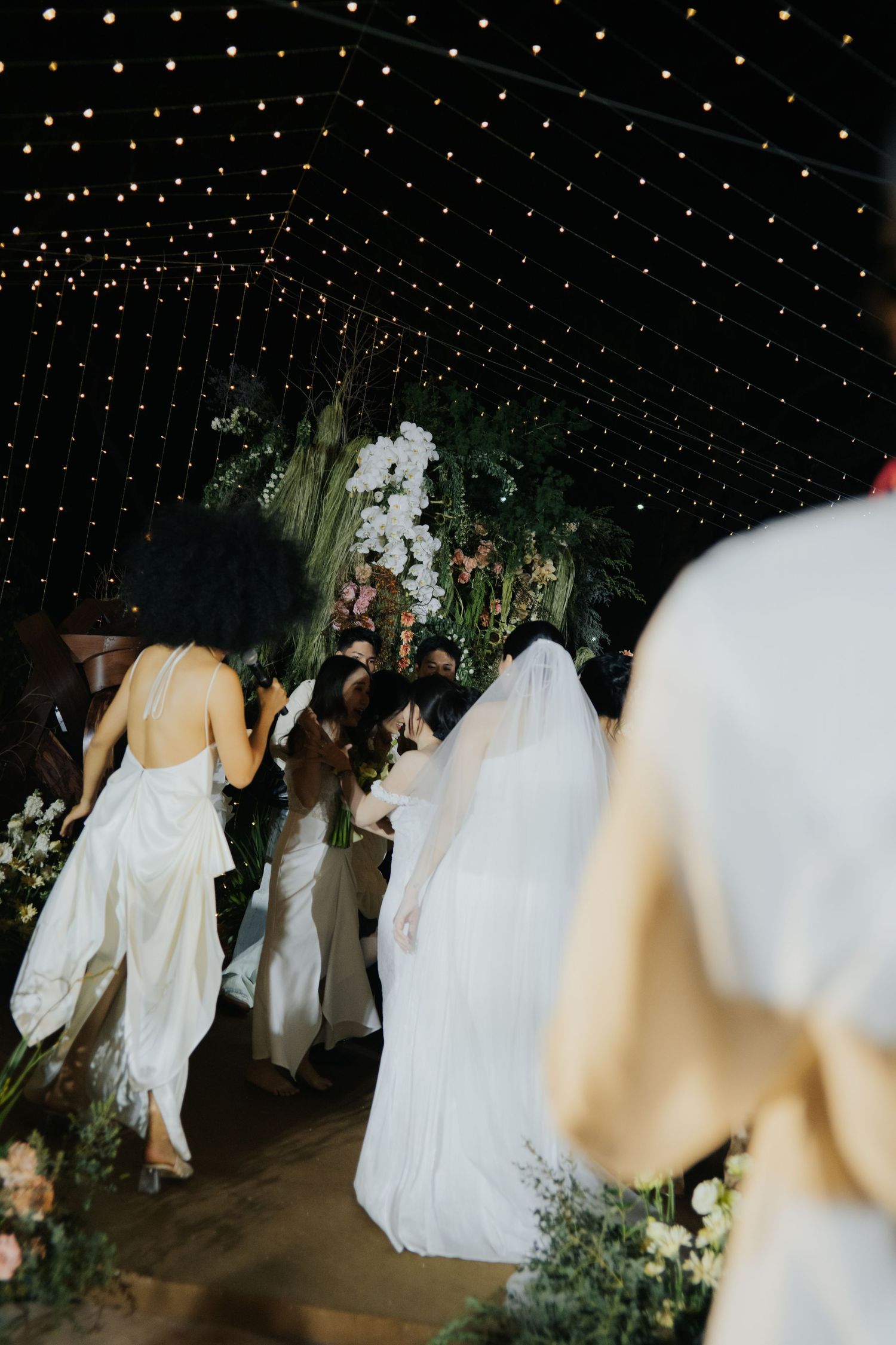 Bride in white gown and veil walks down floral aisle under twinkling fairy lights at elegant wedding.