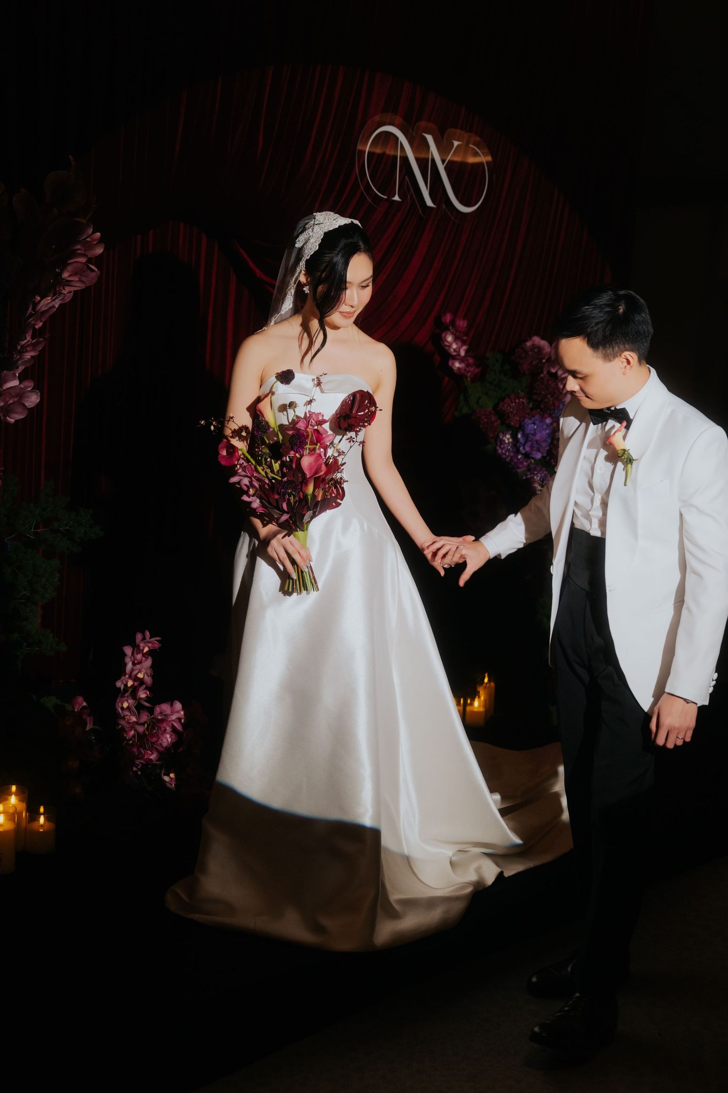 Bride in strapless white gown holds burgundy bouquet as groom in white tuxedo jacket holds her hand at elegant dark wedding.