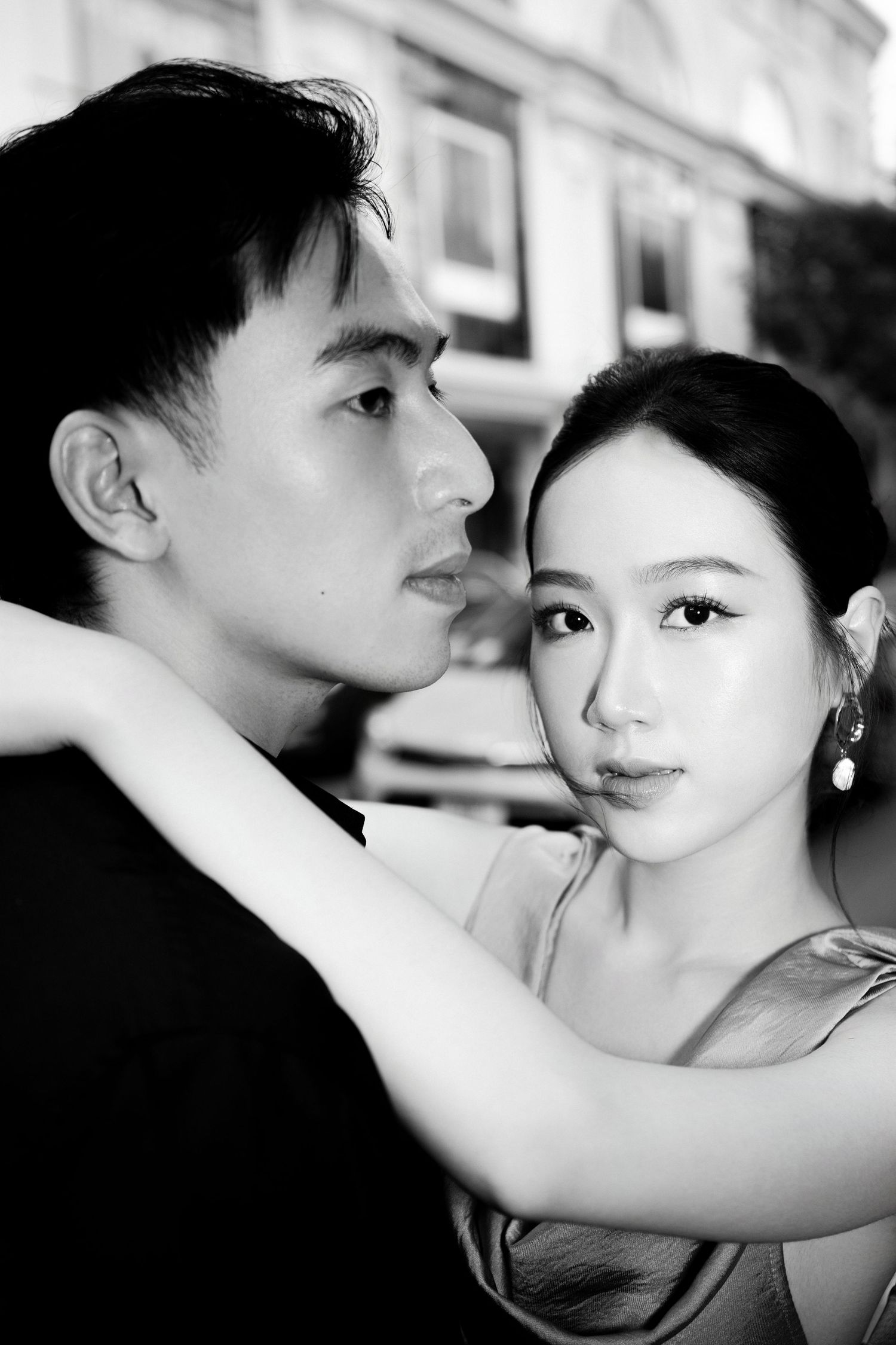 Black and white portrait of an elegant Asian couple embracing closely, woman wearing drop earrings, romantic outdoor setting.