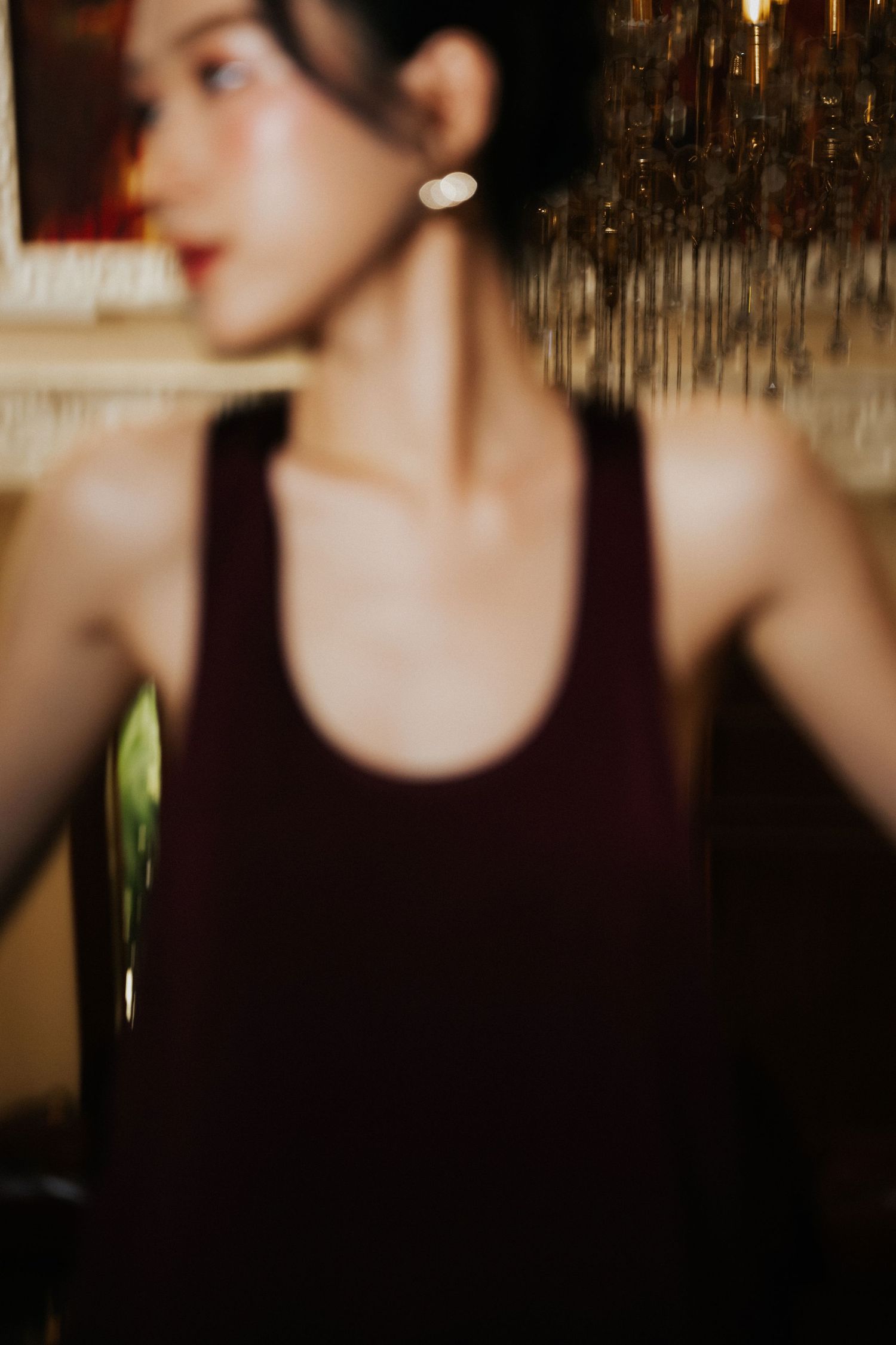 A woman in a dark burgundy tank top with pearl earrings stands in a dimly lit room with bokeh lighting.