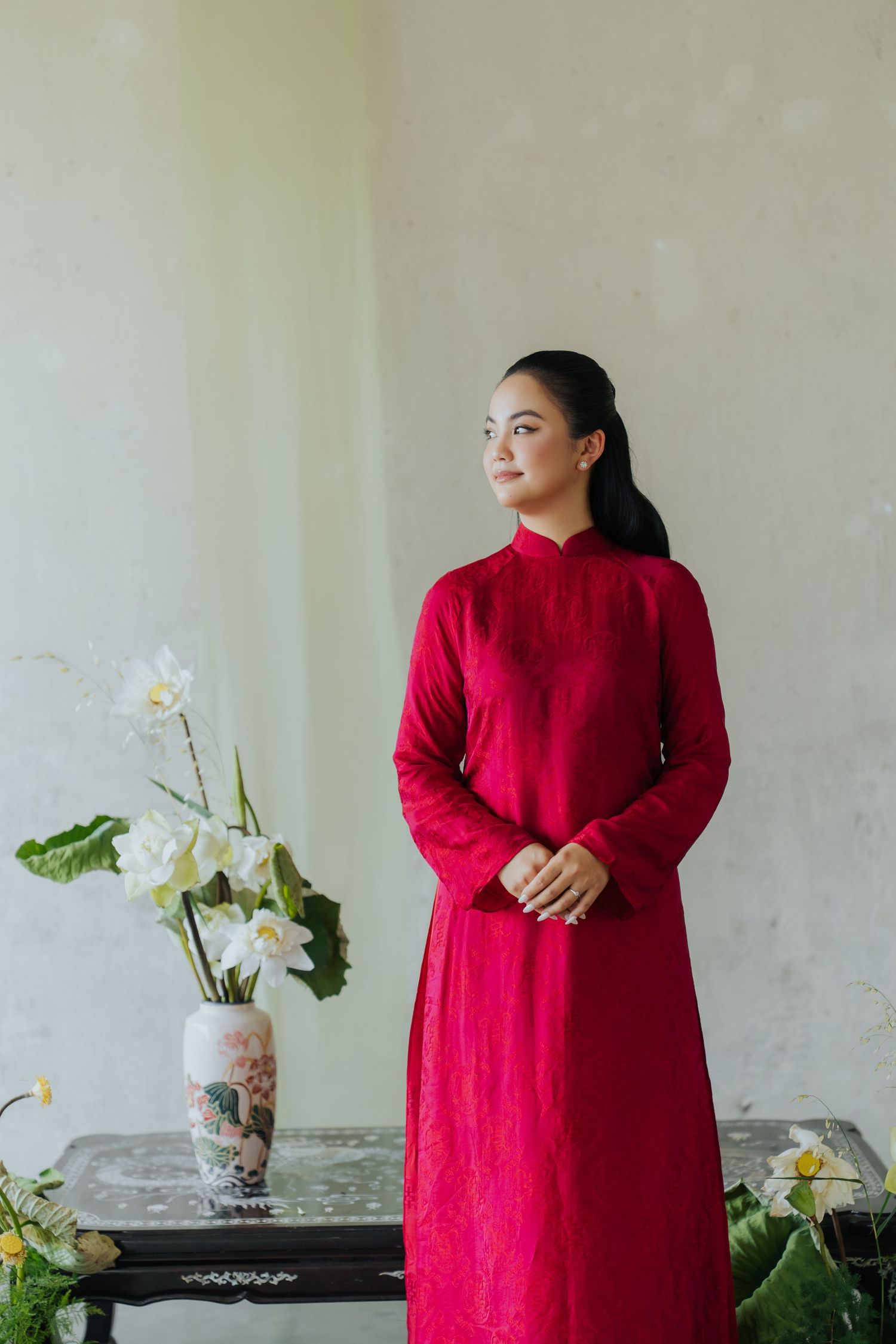 A woman in a red Vietnamese Ao Dai dress stands gracefully beside a vase of white lotus flowers.