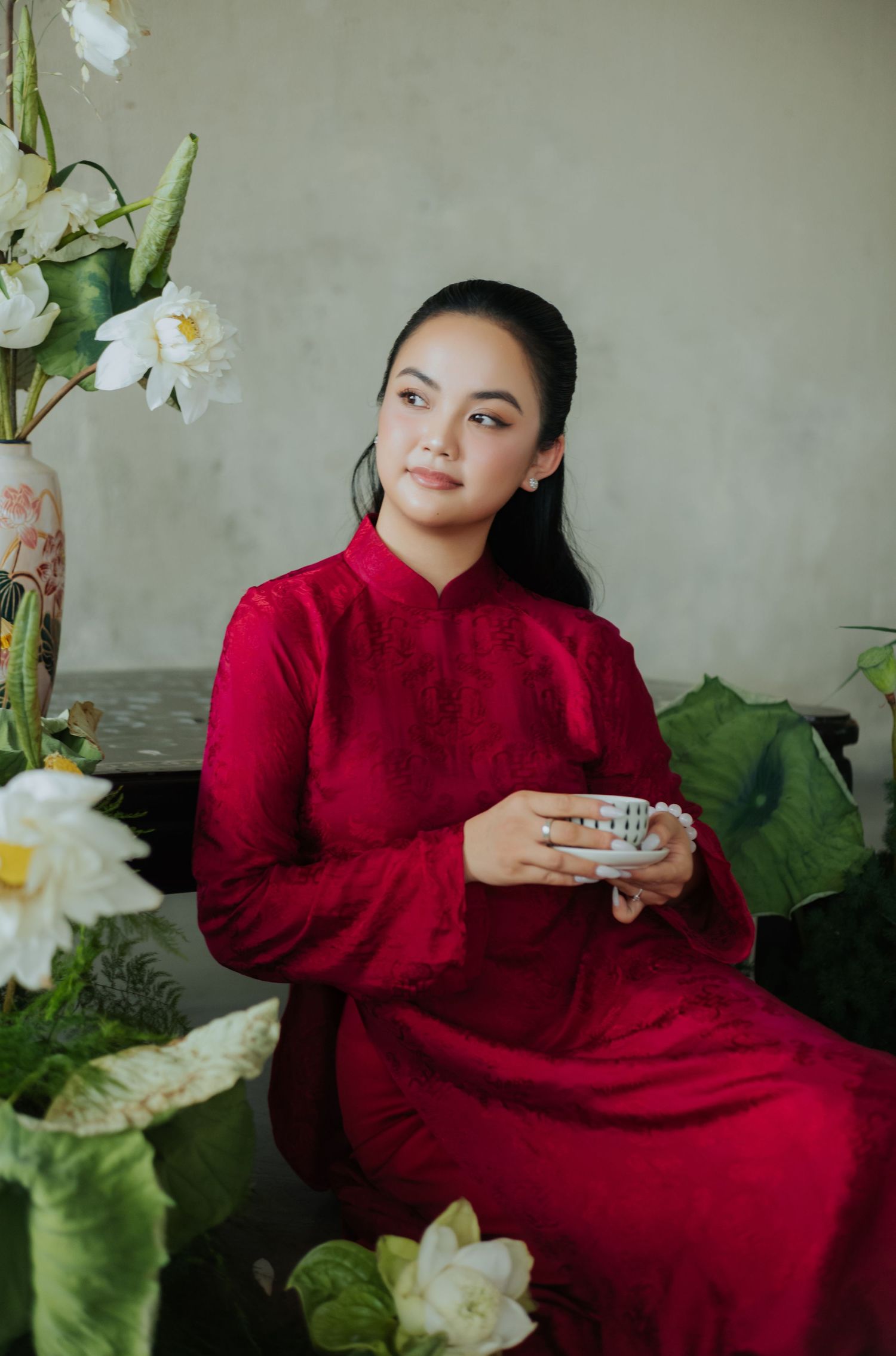 A young woman in a red ao dai holds a teacup, seated among white lotus flowers and green leaves.