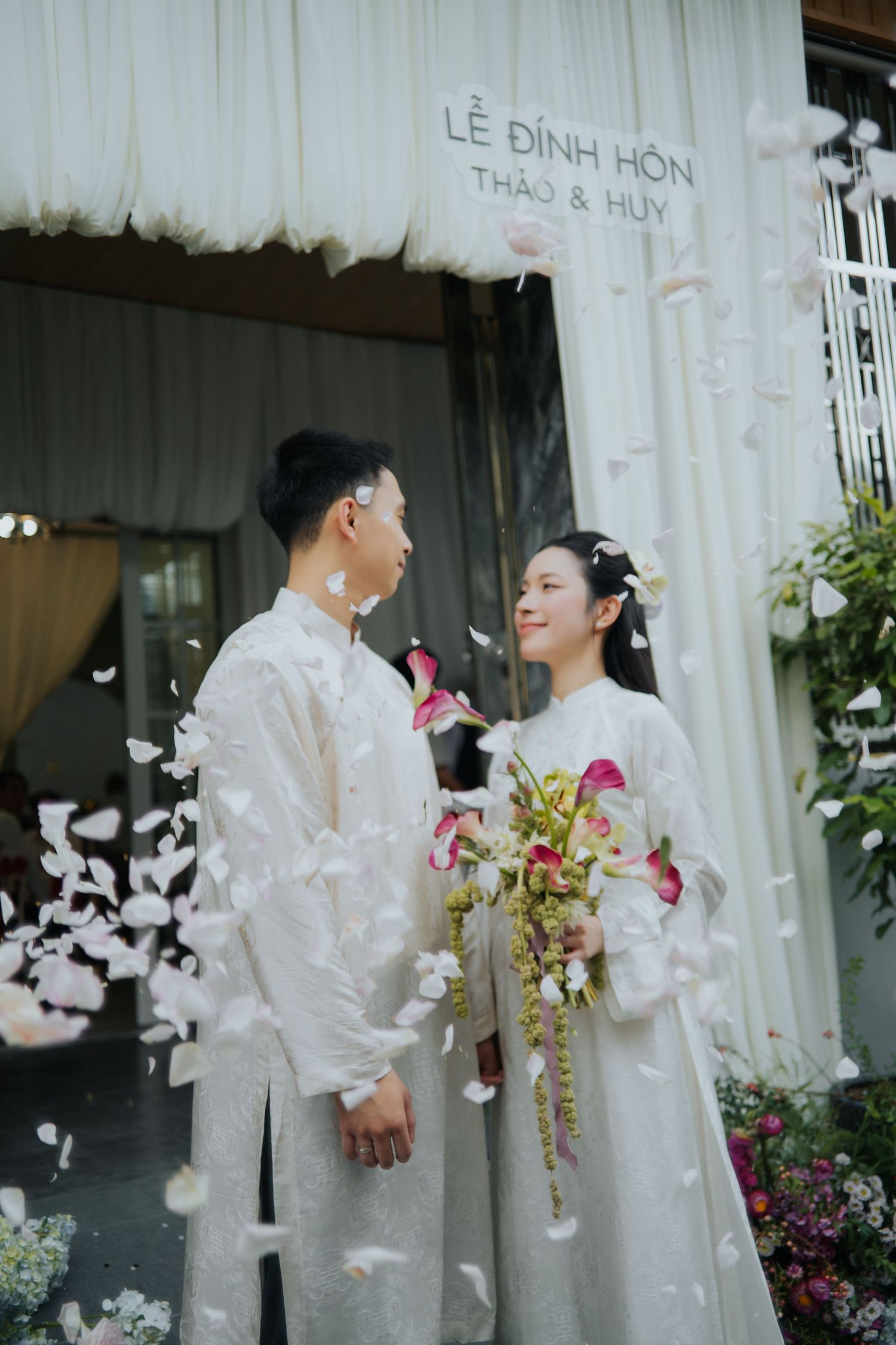 Vietnamese couple in white ao dai attire exchange glances during Le Dinh Hon engagement ceremony with flower petals falling.