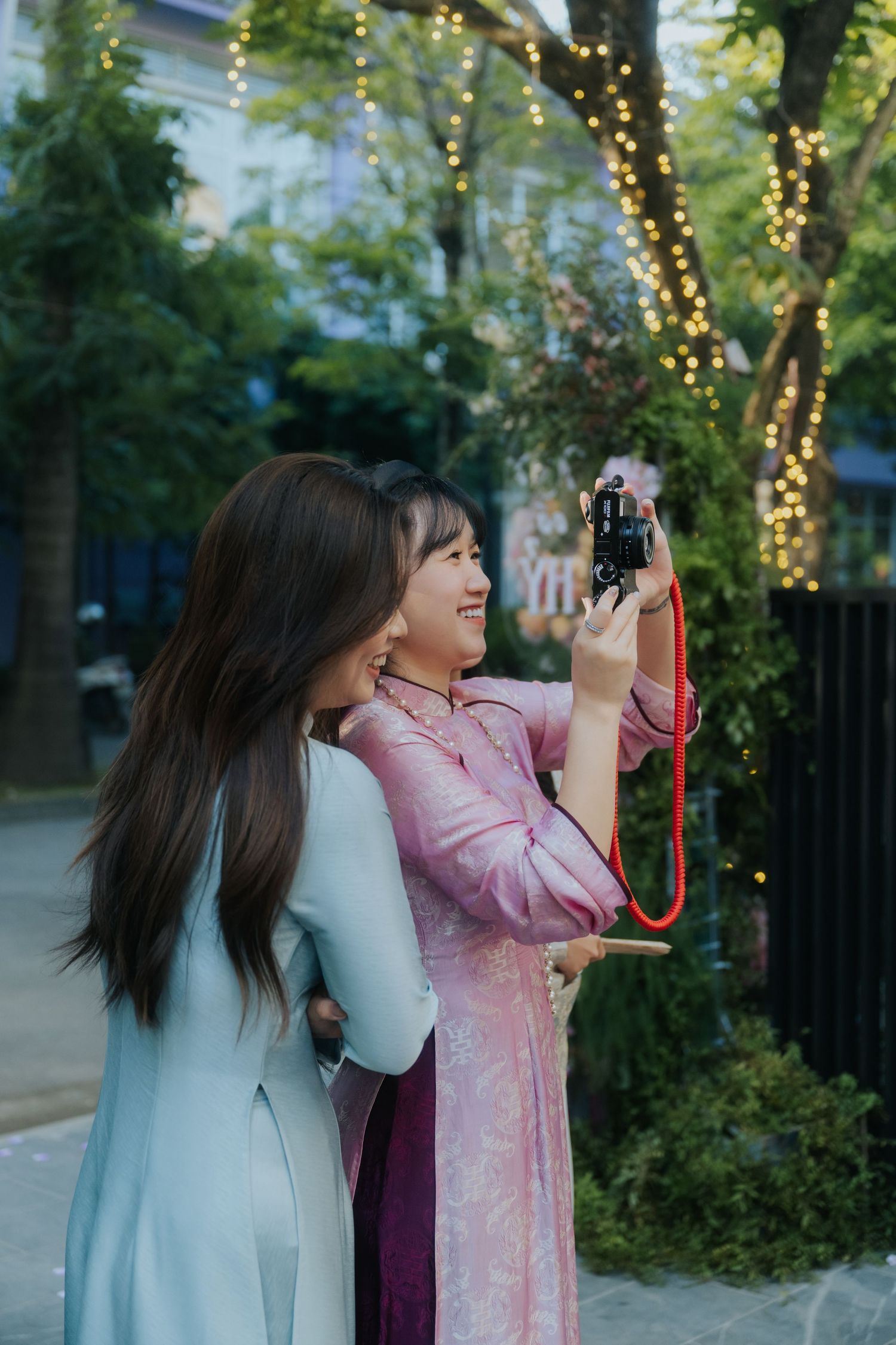 Two young women in colorful Vietnamese ao dai dresses taking photos with a film camera outdoors near fairy lights.