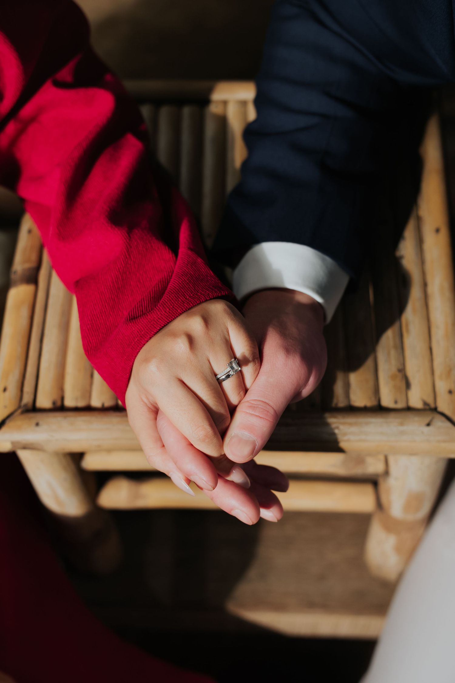 Couple holding hands on bamboo chair, woman wearing diamond engagement ring, red sleeve and navy suit visible.