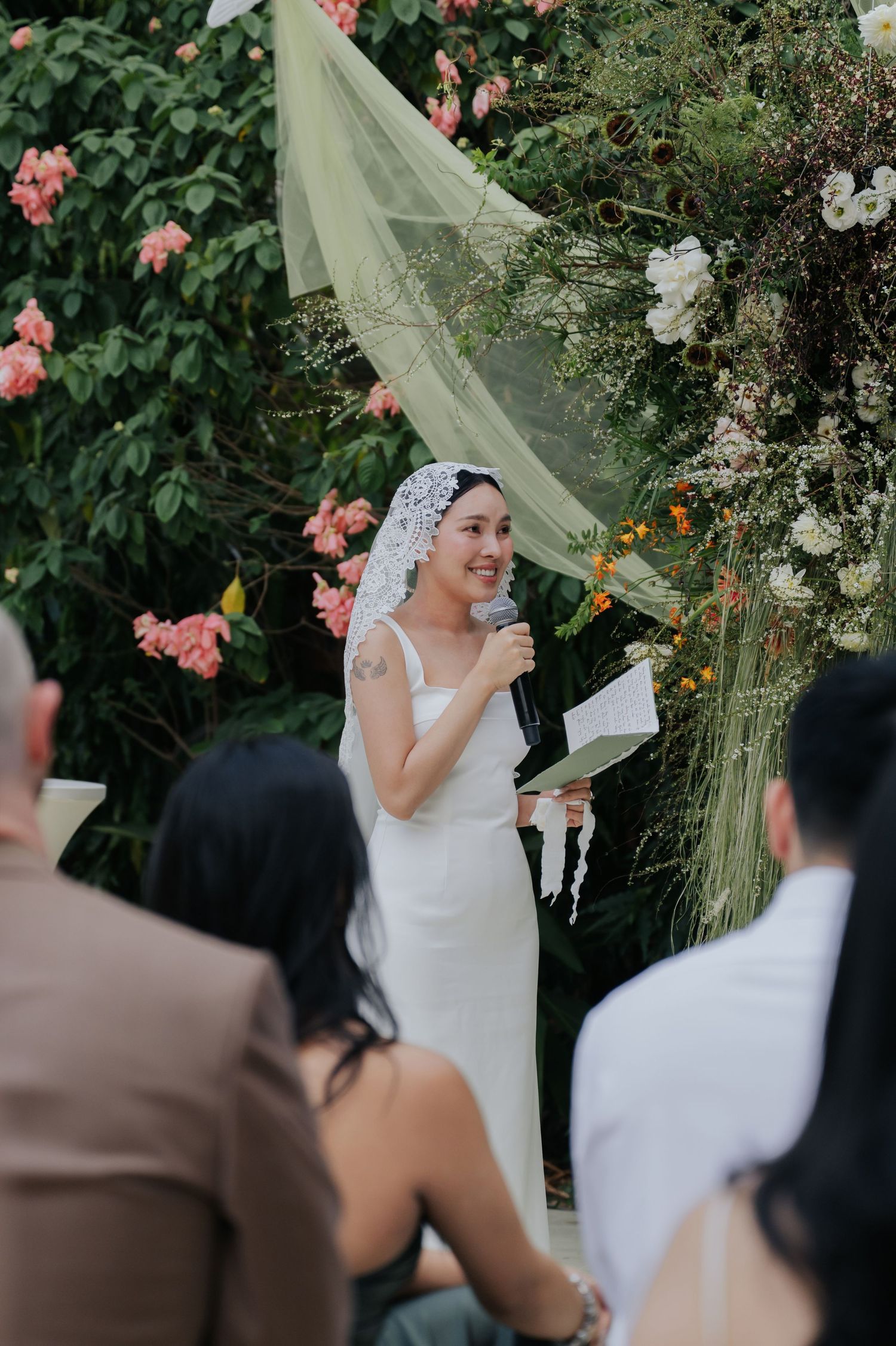 Bride in white dress and veil speaks at outdoor garden wedding ceremony with floral arch backdrop.