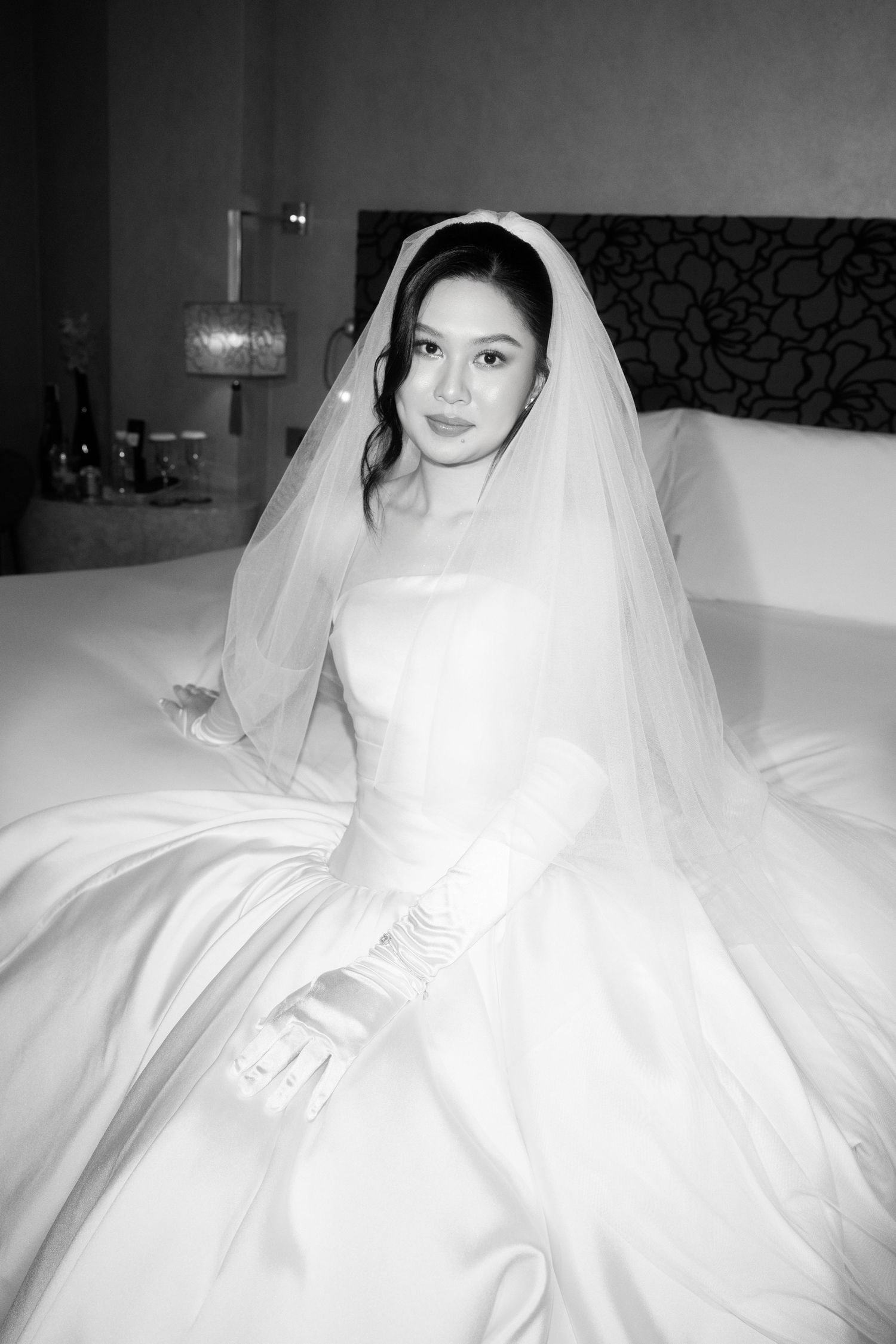 Black and white photo of a bride in a white gown, veil, and gloves sitting elegantly on a bed.