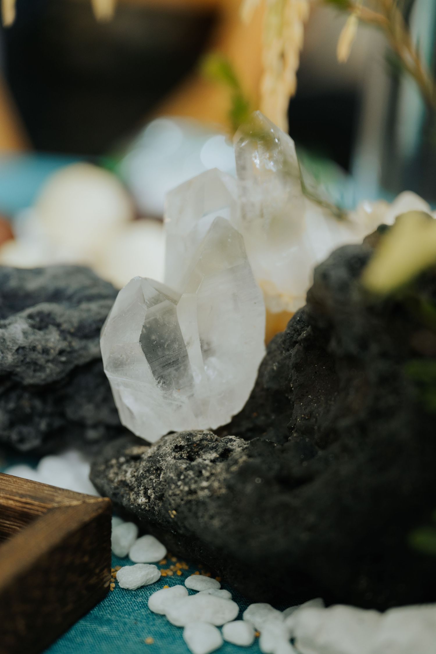 Clear quartz crystal cluster resting on dark volcanic rock surrounded by white pebbles and minerals on a teal surface.