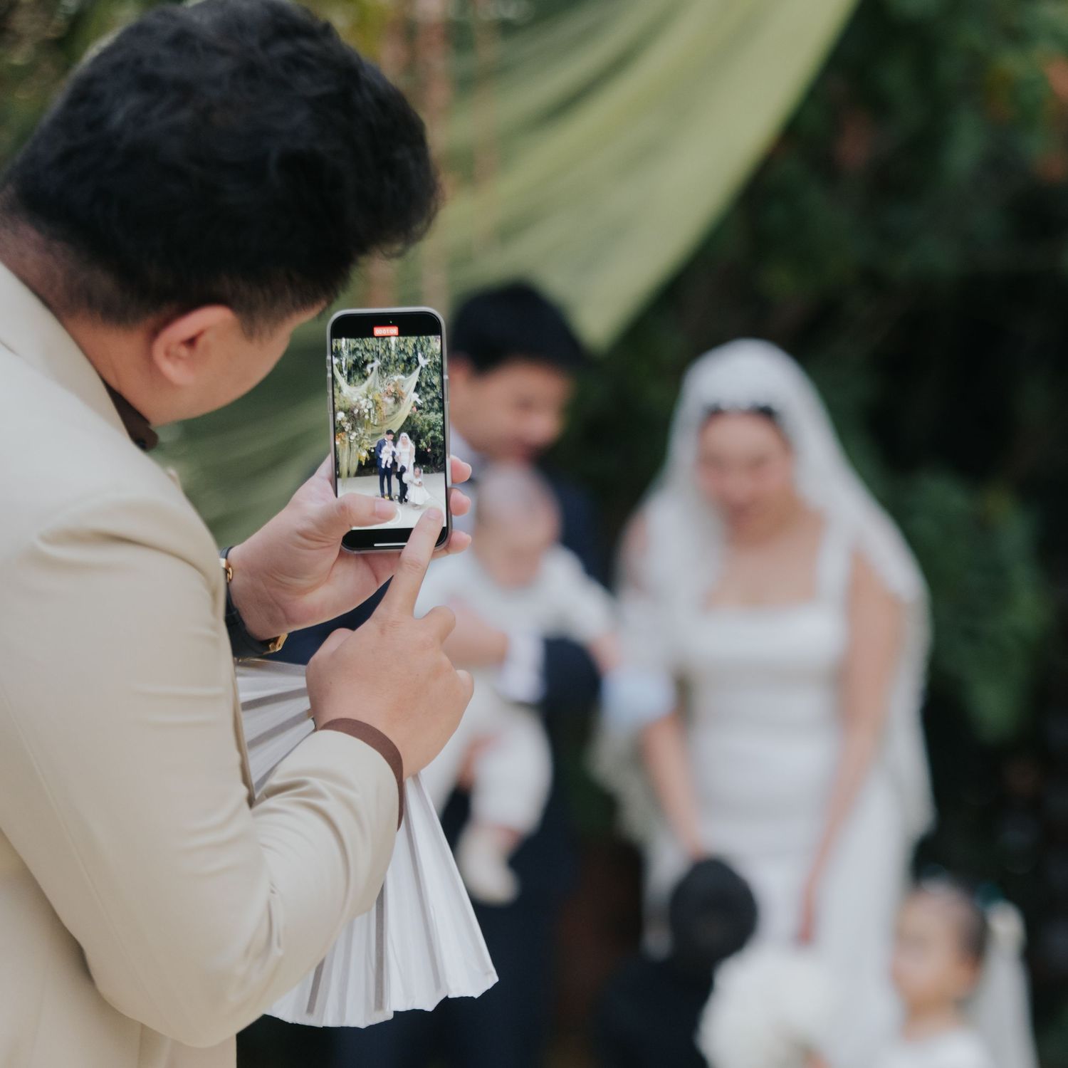 A man in a beige suit photographs a bride and groom at an outdoor wedding ceremony using a smartphone.