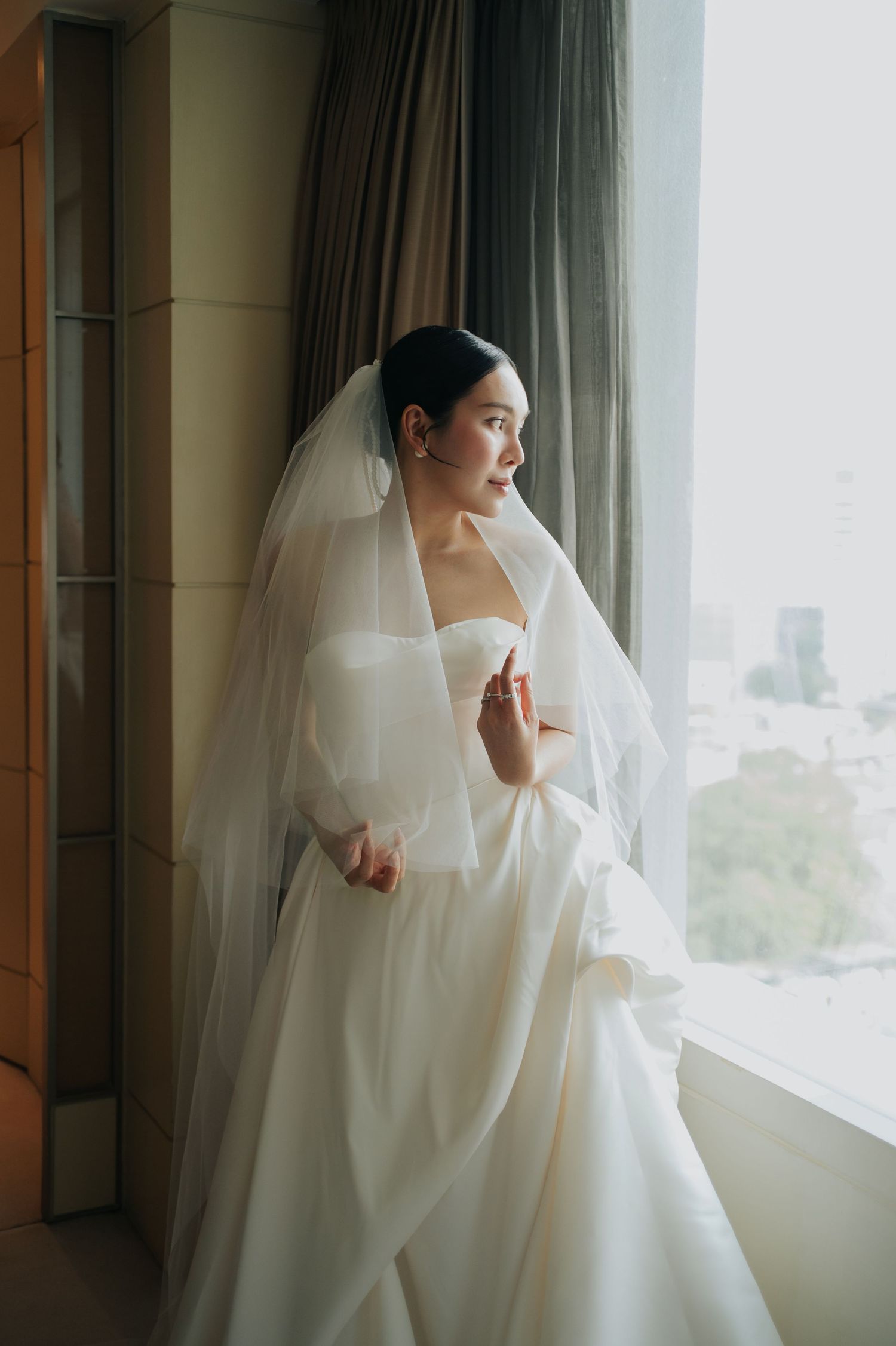 Bride in strapless white ball gown and veil gazes out a bright window, holding her dress elegantly.