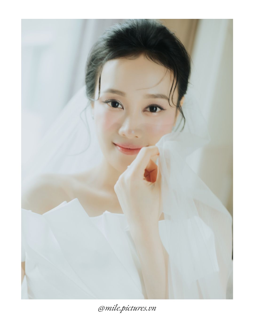 A beautiful Asian bride in a white strapless gown and veil, posing elegantly with hand near face.