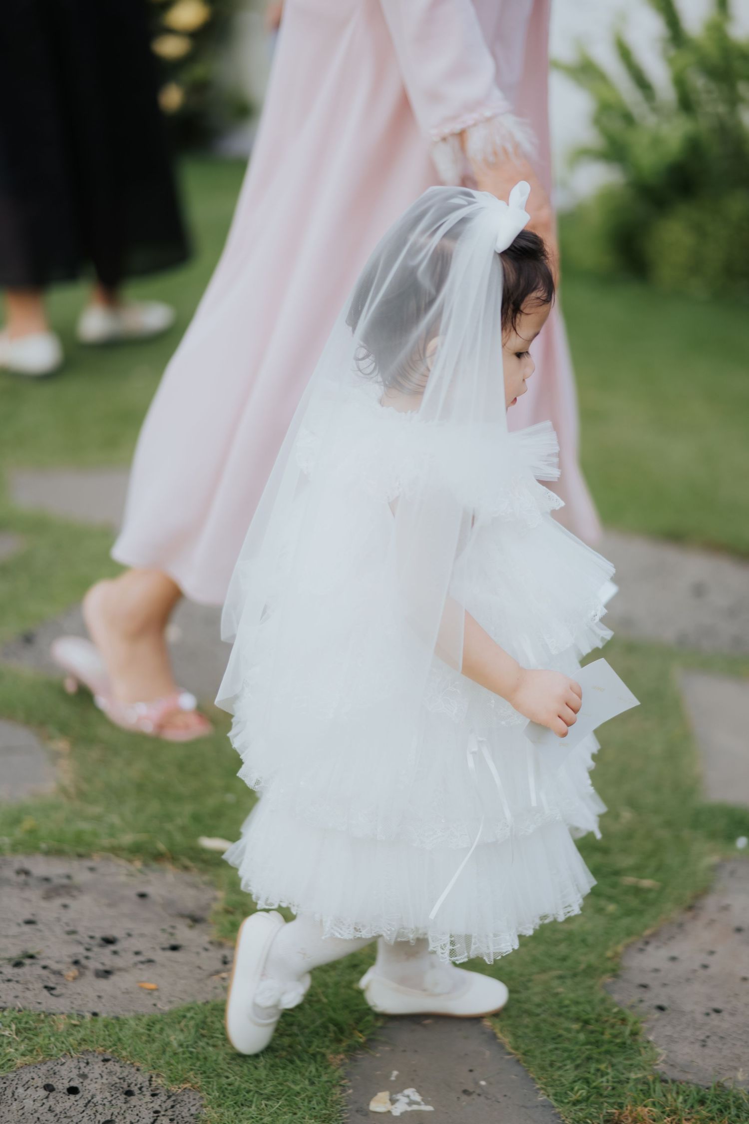 Adorable flower girl in white ruffled dress and veil walks beside woman in pink gown at outdoor wedding.