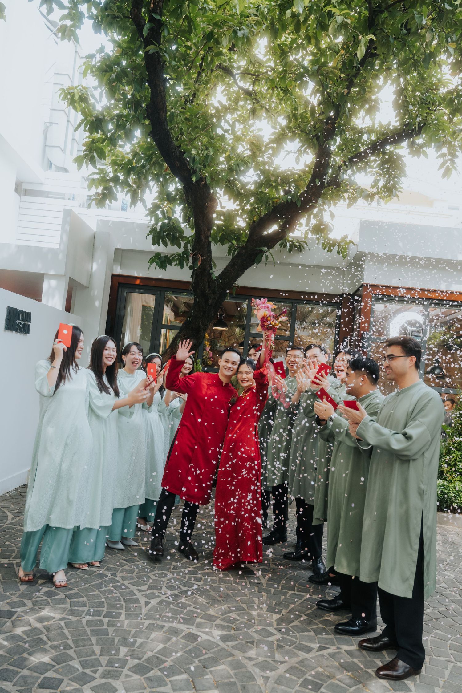 Vietnamese wedding ceremony with couple in red ao dai, surrounded by guests in green traditional attire, celebrating with confetti.
