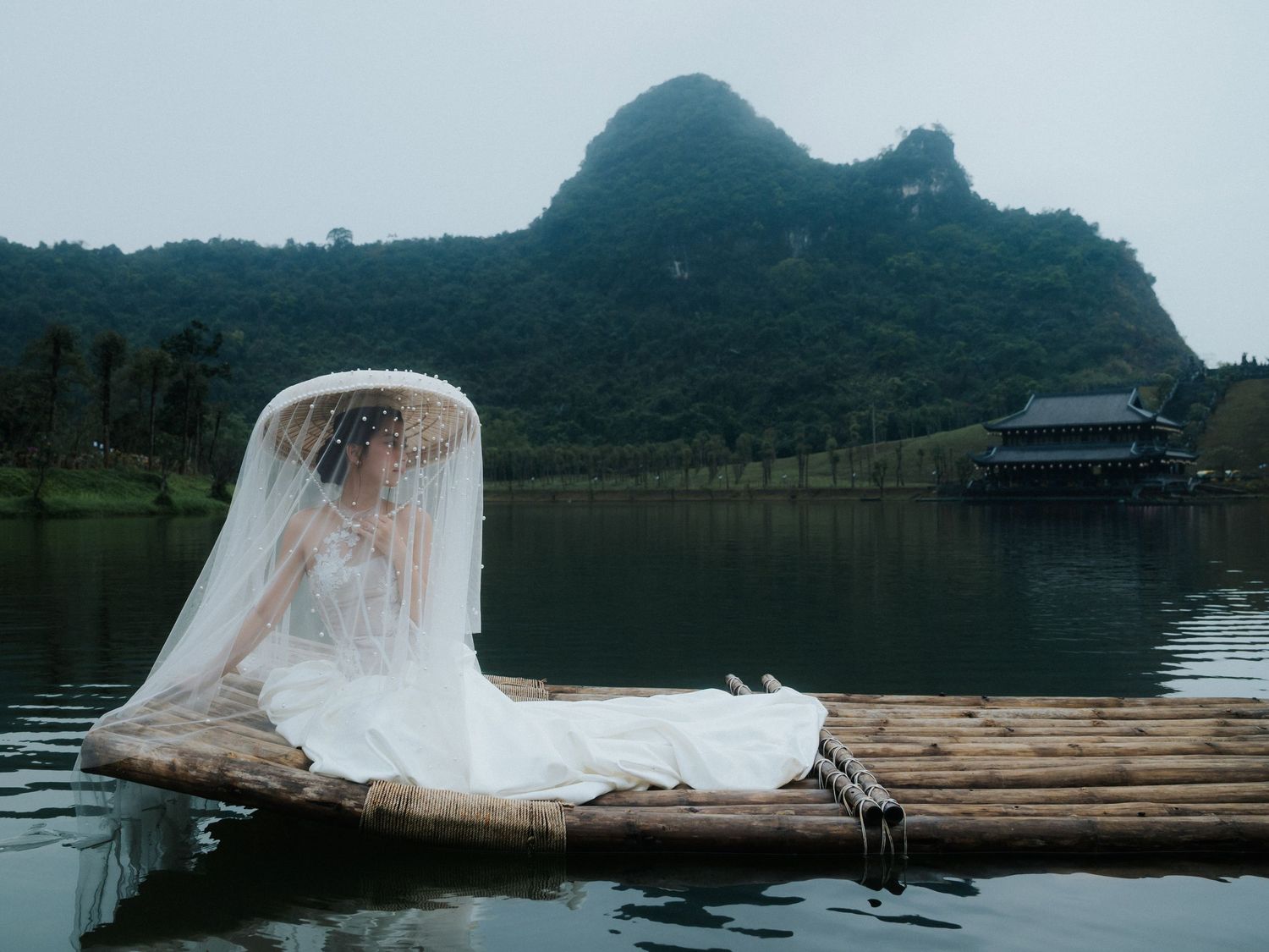 Bride in white gown and veil sits on bamboo raft on calm lake with karst mountains and pagoda backdrop.