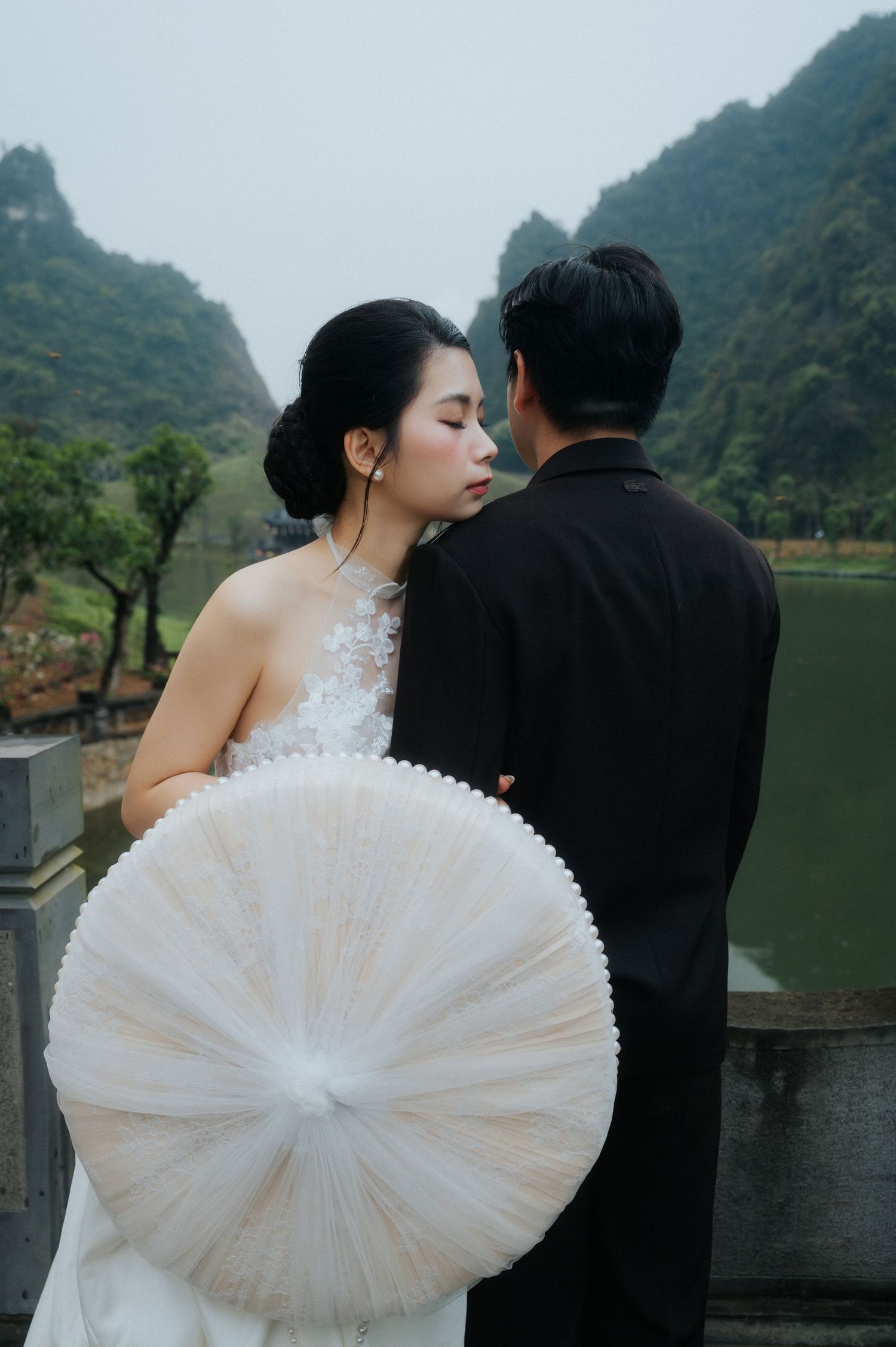 A bride in a white lace dress holds a parasol while standing with a groom in black near a misty mountain lake.