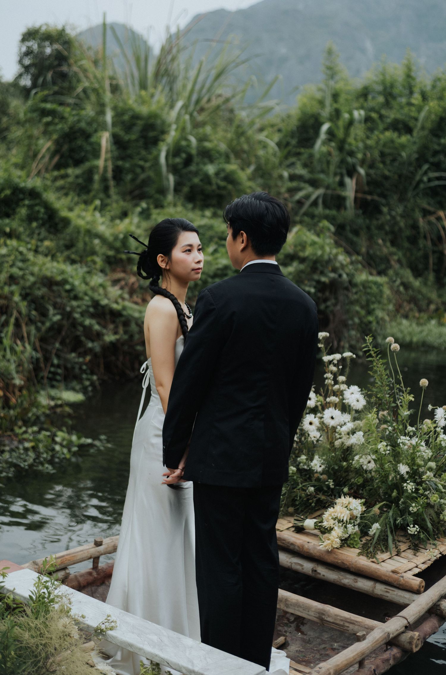 Couple in wedding attire standing on a bamboo raft surrounded by lush greenery and white flowers by a serene river.