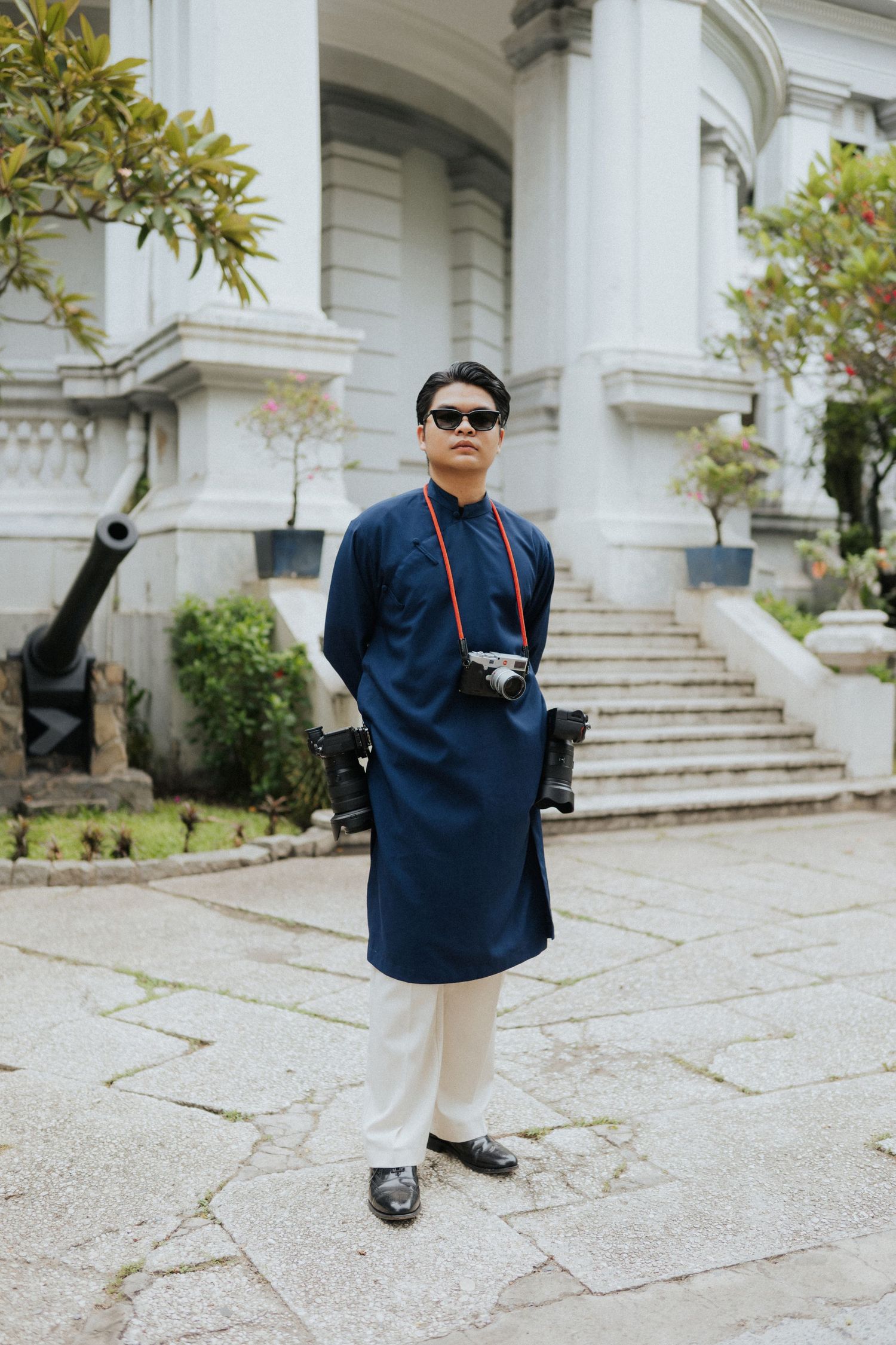 Man in navy blue ao dai traditional Vietnamese outfit with sunglasses and camera standing before white colonial building.