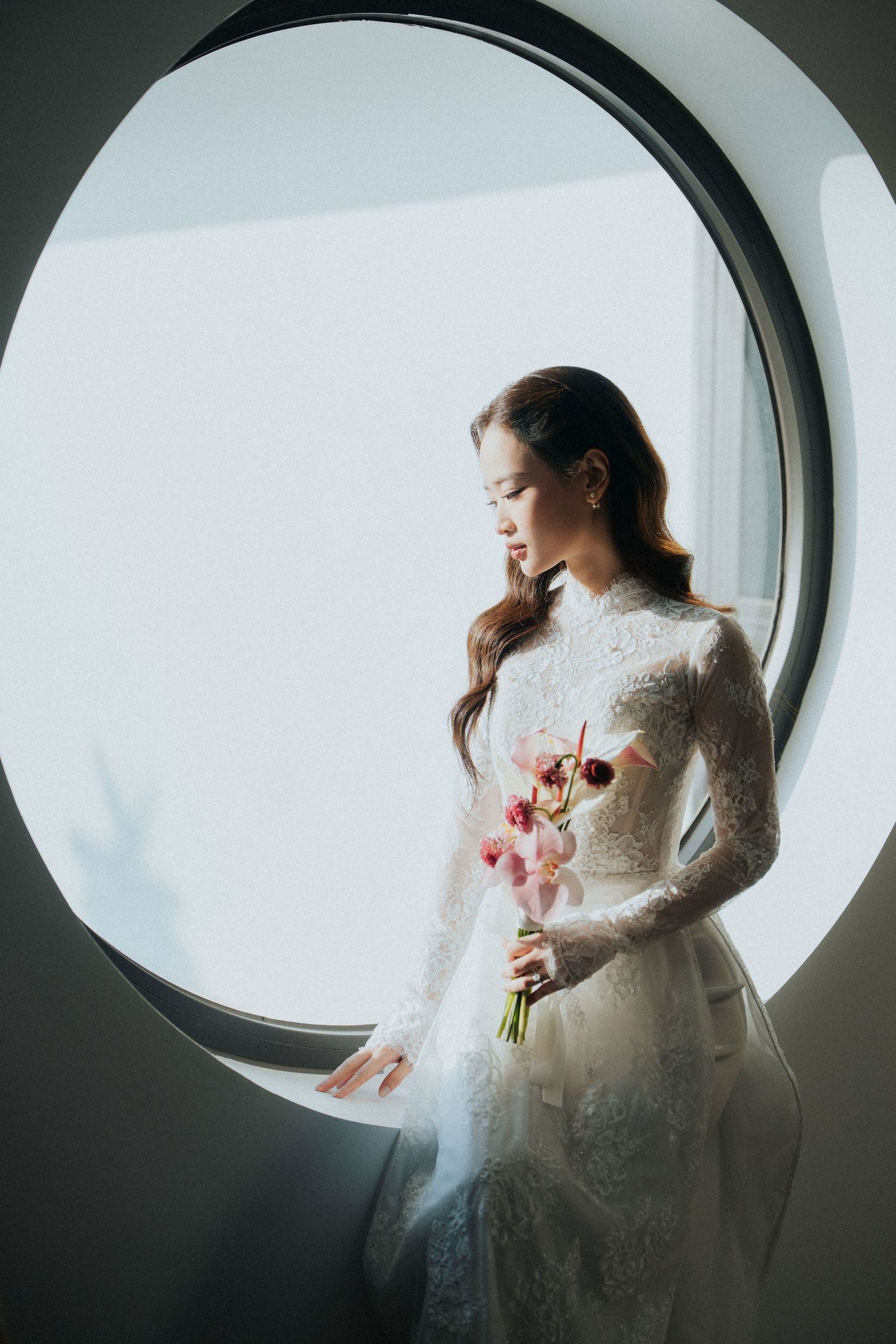 Bride in white lace gown holding pink flowers stands by circular window in soft natural light.
