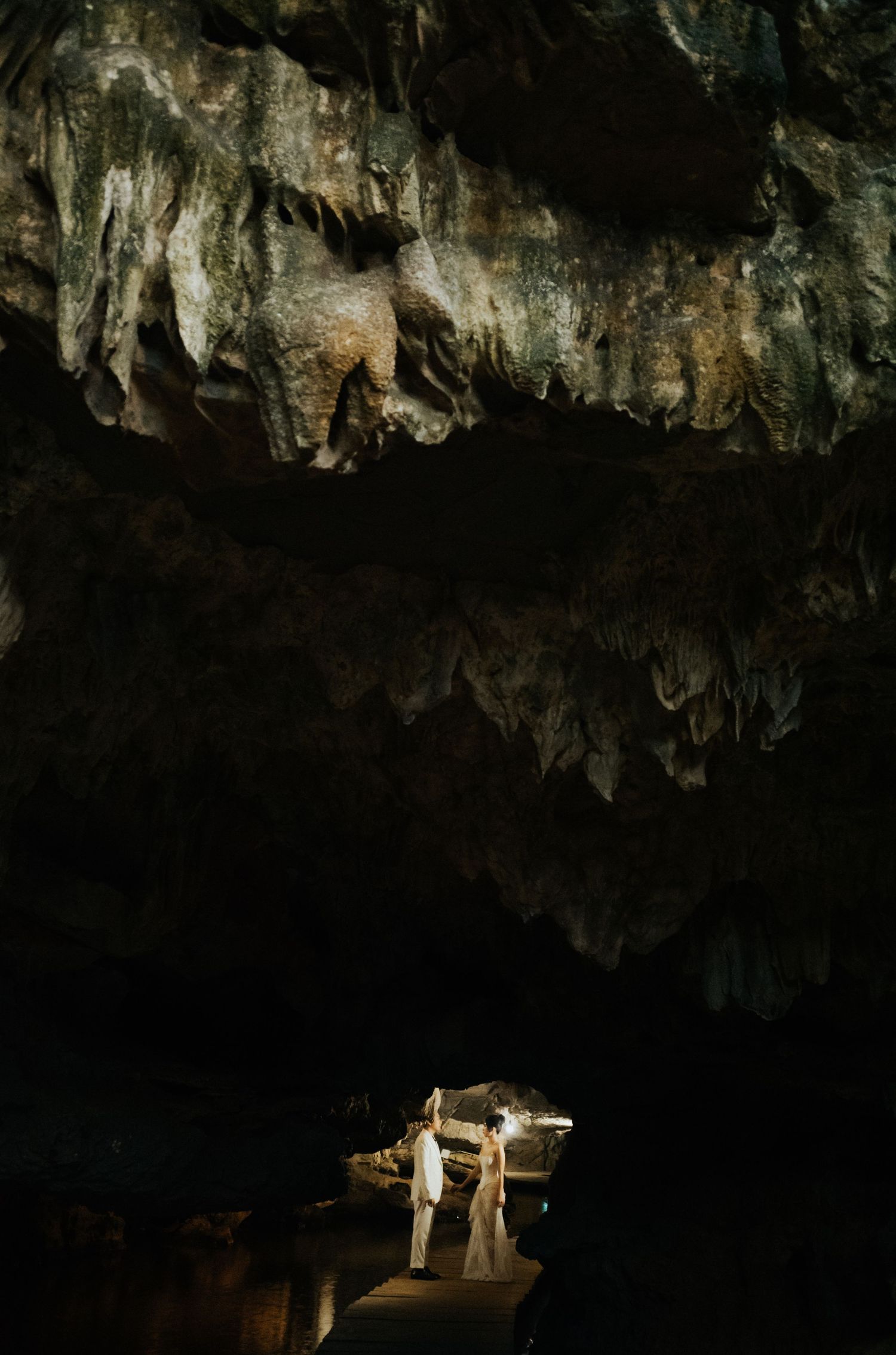 Couple in white attire standing in a dramatic cave with stalactites and natural rock formations illuminated by soft light.