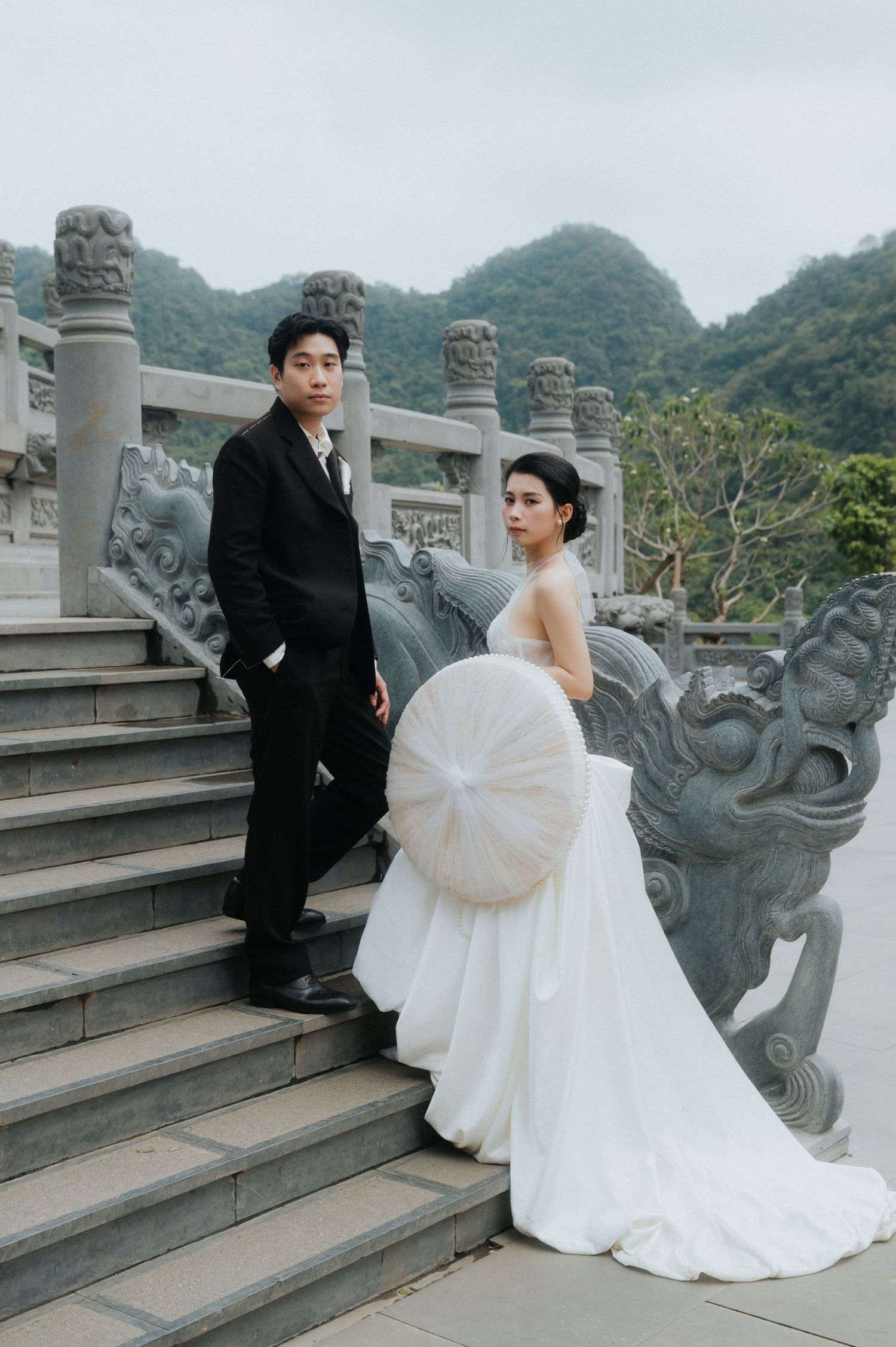Couple in wedding attire poses on ornate stone steps with dragon sculptures and misty mountains in background.