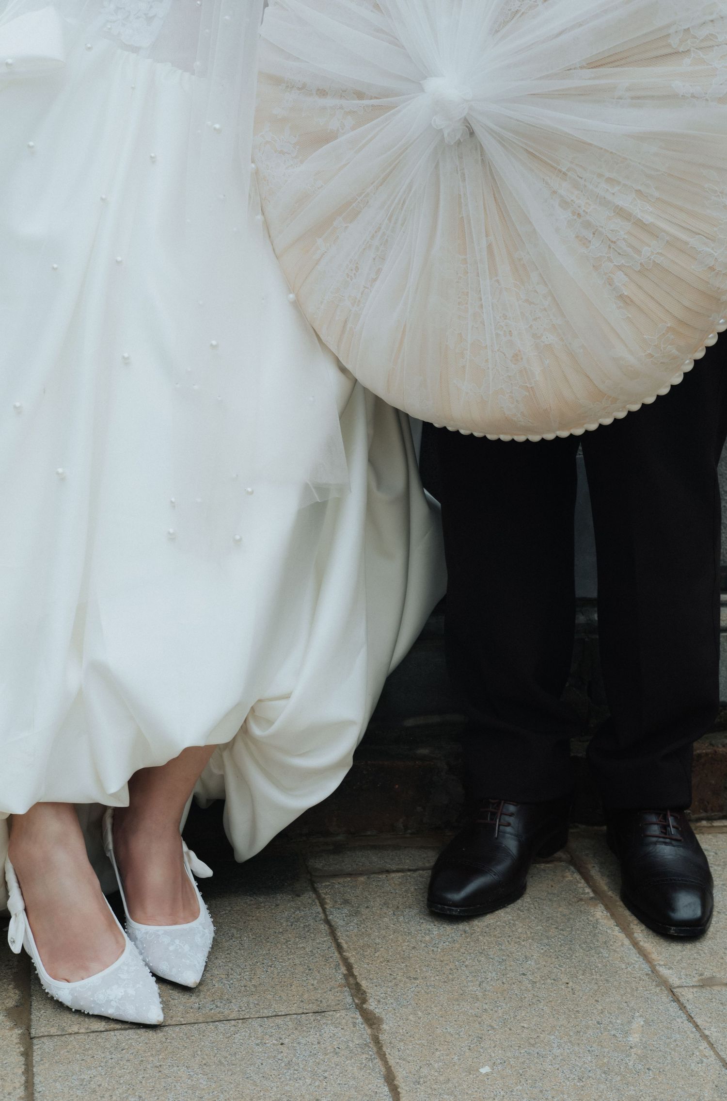 Bride in white wedding dress and lace heels stands beside groom in black suit holding a traditional parasol umbrella.