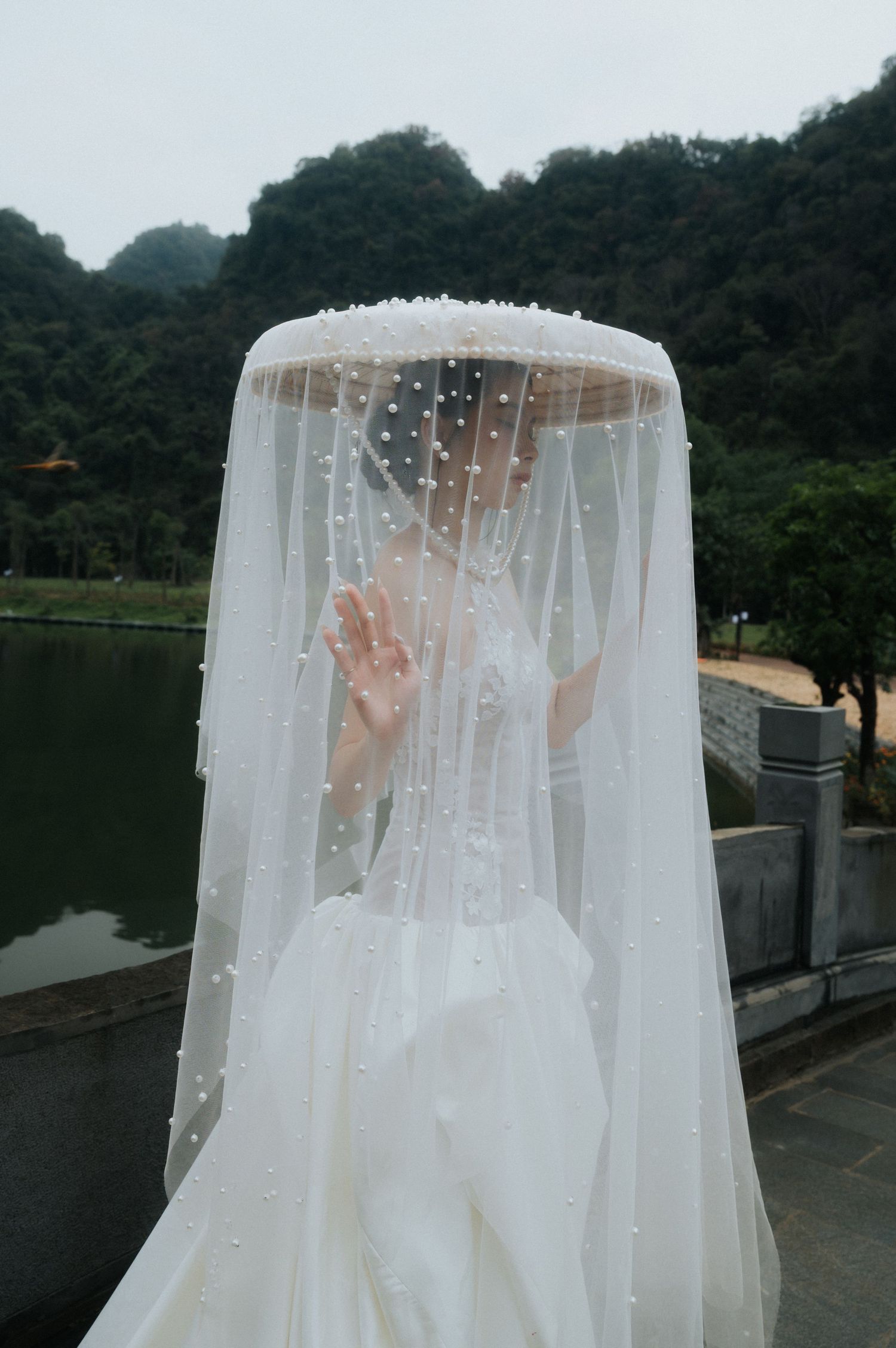 Bride in white wedding dress and pearl-adorned veil covering face, standing by a lake with green mountains backdrop.