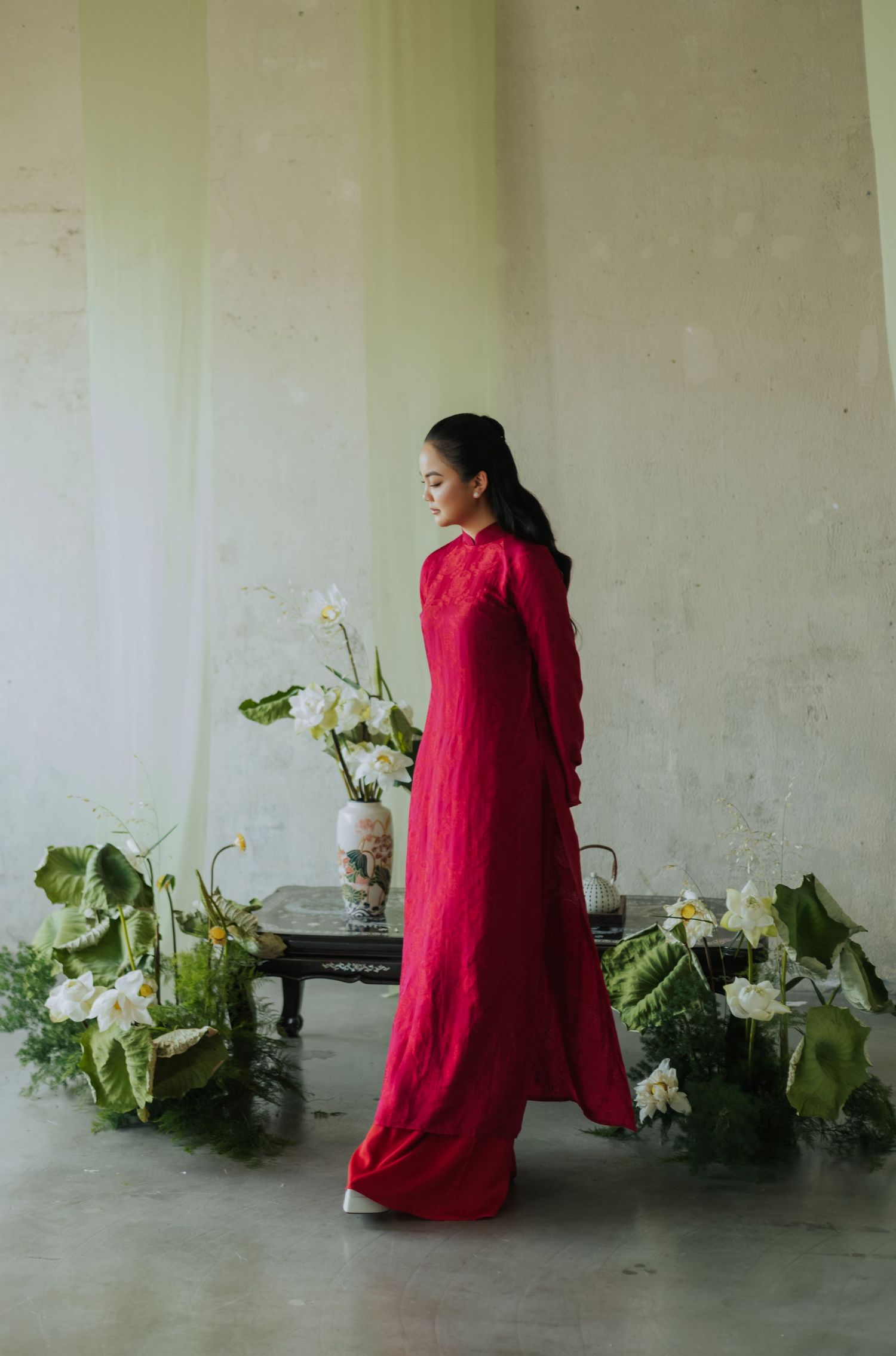 Woman in elegant red maxi dress standing near floral arrangement with white flowers and green leaves.