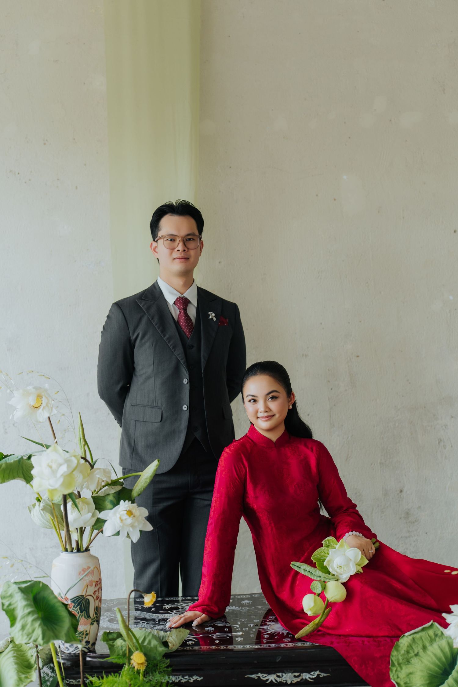 A couple in traditional Vietnamese attire, man in dark suit and woman in red ao dai, posing elegantly with white flowers.