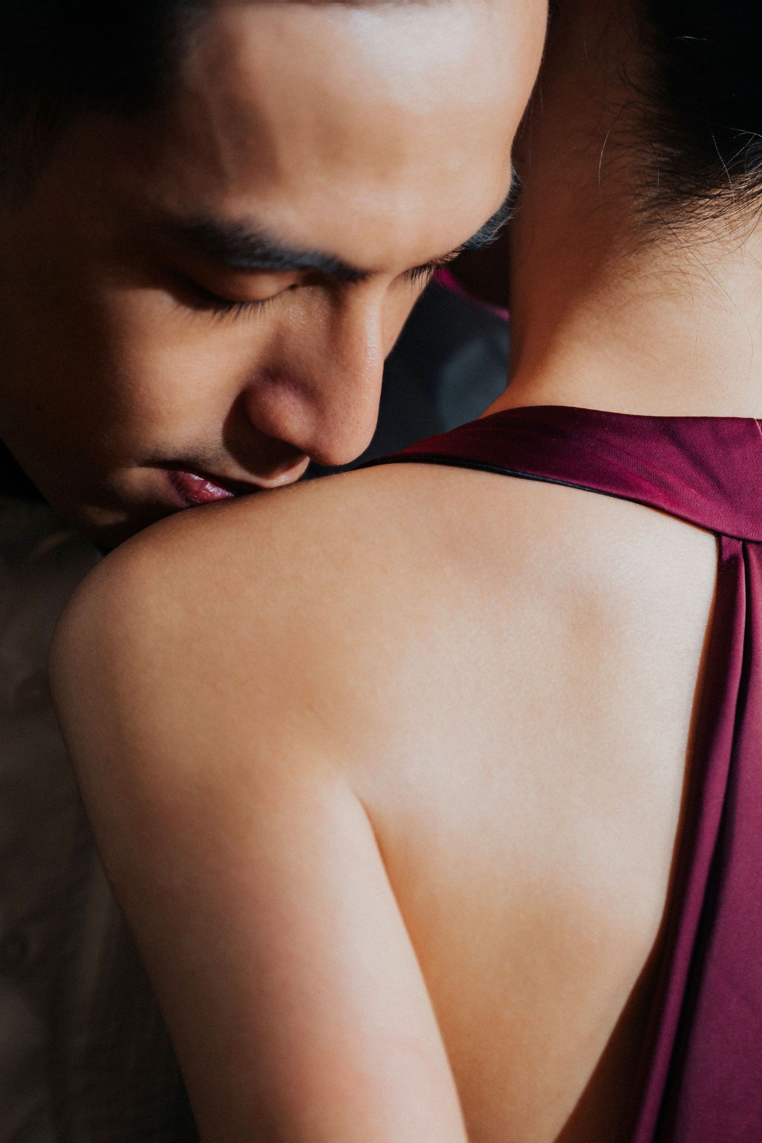 A man gently kisses a woman's shoulder as she faces away in a burgundy dress.
