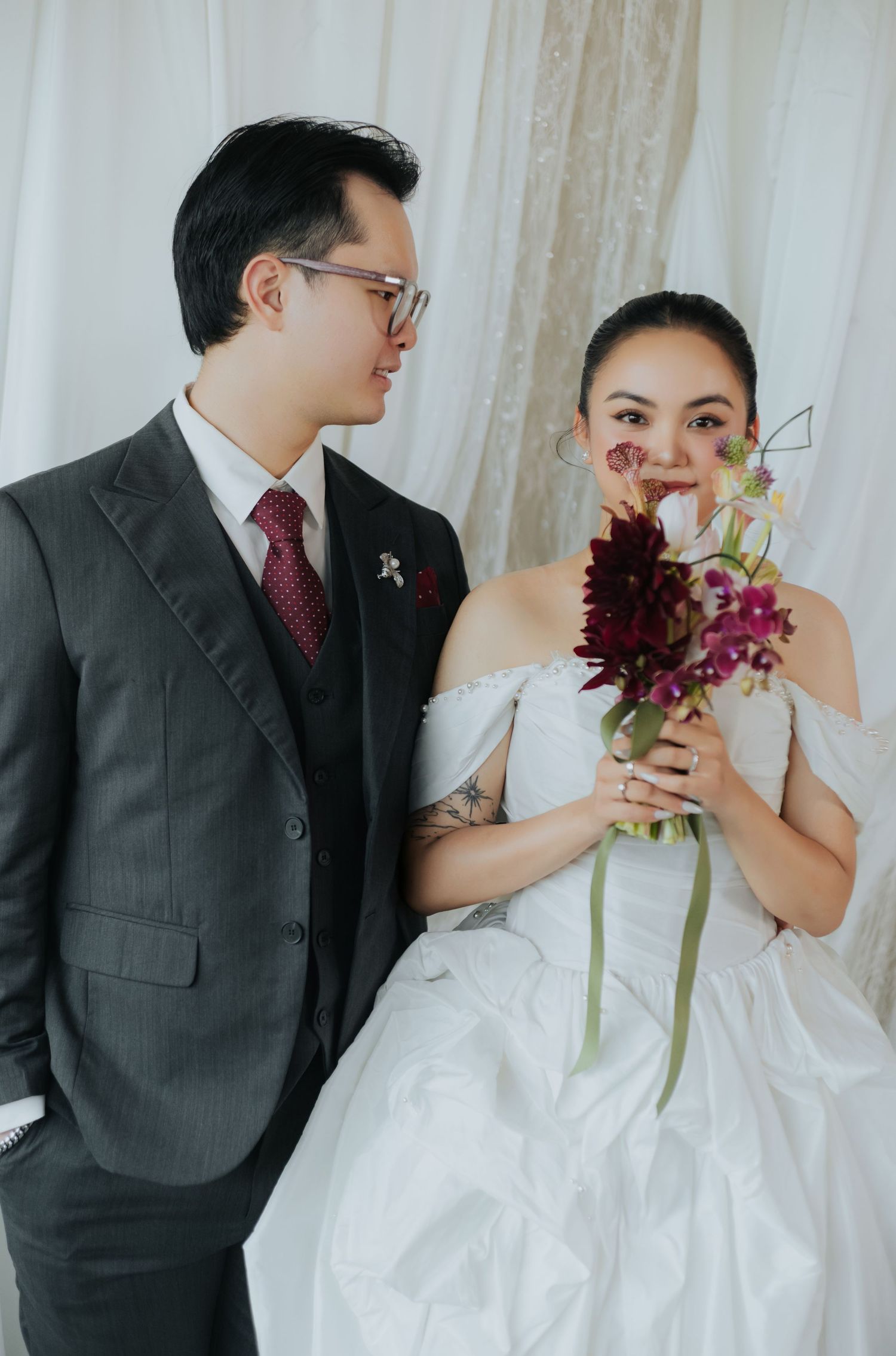 A bride in a white off-shoulder gown holds a bouquet while a groom in a gray suit with red tie gazes at her lovingly.