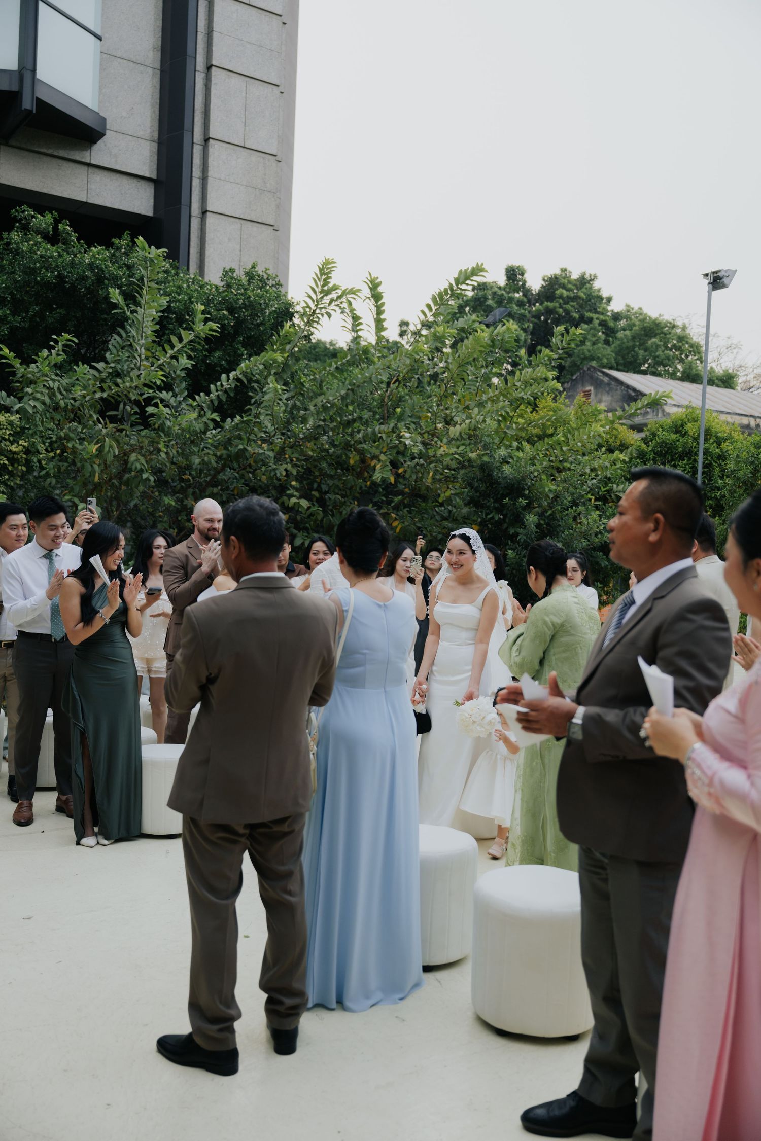 Outdoor wedding ceremony with guests applauding as bride in white gown walks through an aisle surrounded by lush greenery.