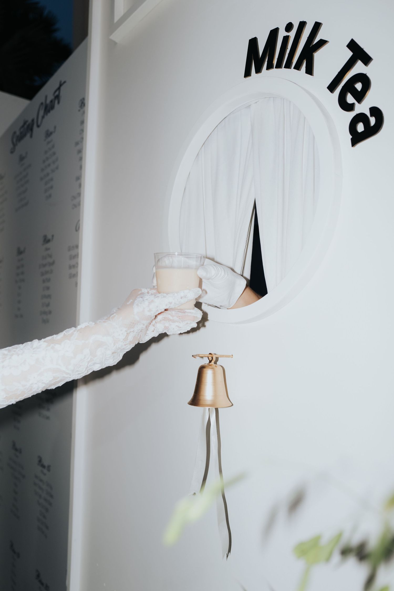 A gloved hand holds a milk tea glass through a circular window with a gold bell hanging below a Milk Tea sign.