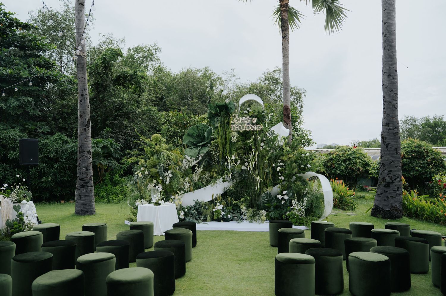 Outdoor wedding ceremony setup with green velvet cylinder seats, floral arch, and tropical palm trees on a lush lawn.