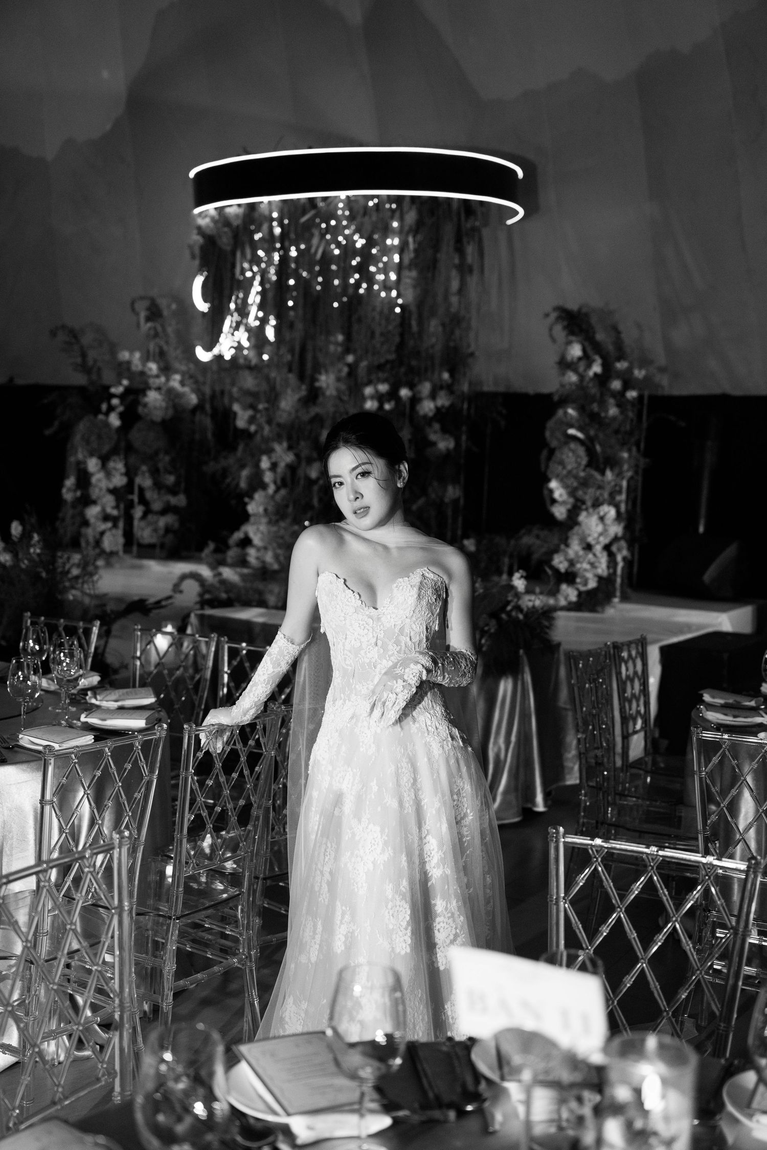 Black and white photo of a bride in a strapless lace gown standing in an elegantly decorated wedding reception venue.