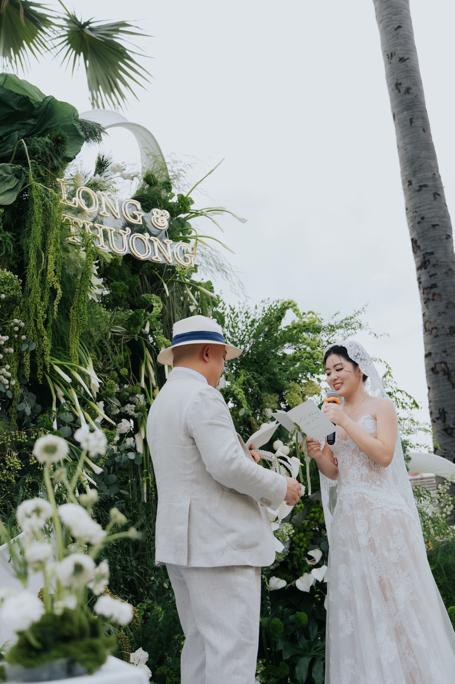 Couple exchanging vows at outdoor garden wedding ceremony with lush greenery backdrop and elegant white floral arrangements.