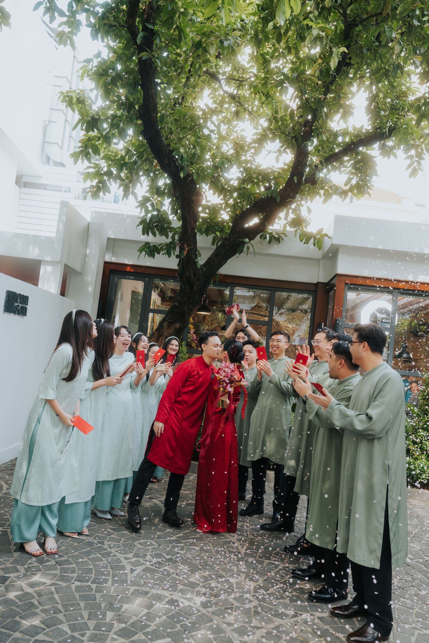 Couple in red ao dai surrounded by guests in sage green traditional Vietnamese attire celebrating outdoors under a lush tree.