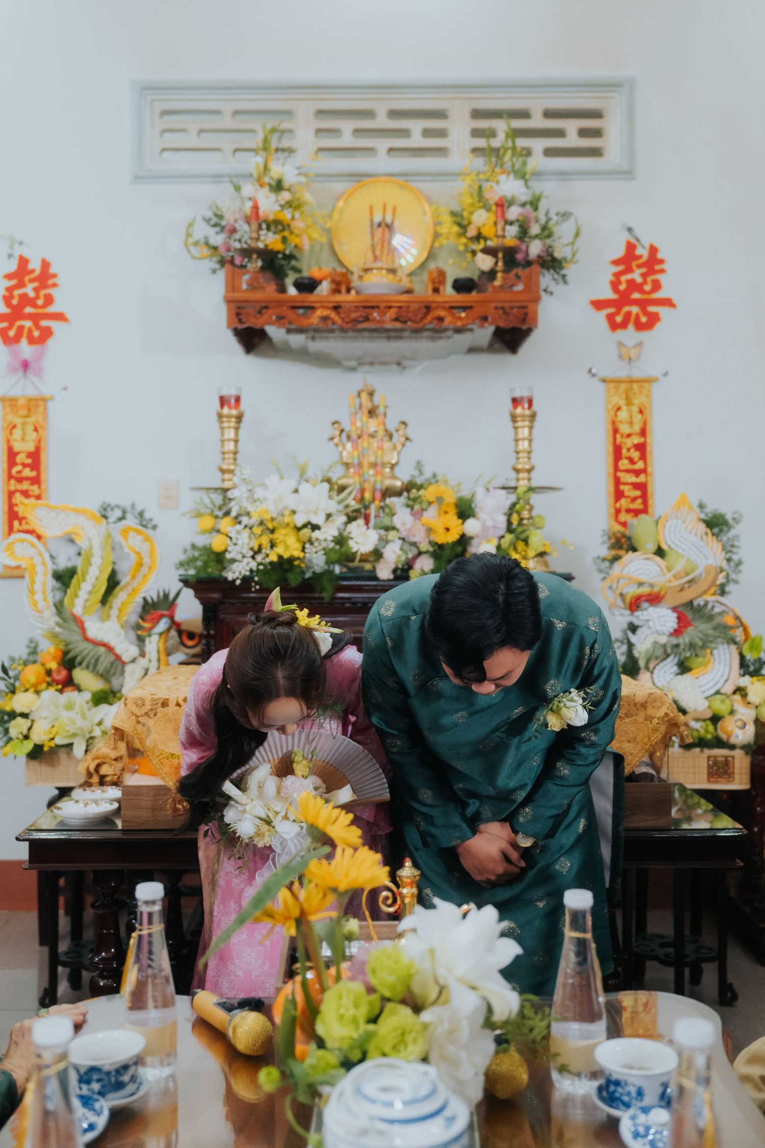 Couple bowing respectfully at a traditional Vietnamese altar adorned with flowers and gold decorations during a ceremony.