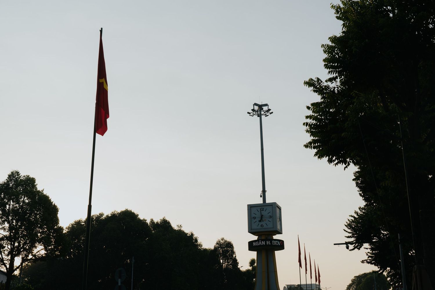 Vietnamese flag on tall flagpole at dusk with silhouetted trees and street light against clear sky.