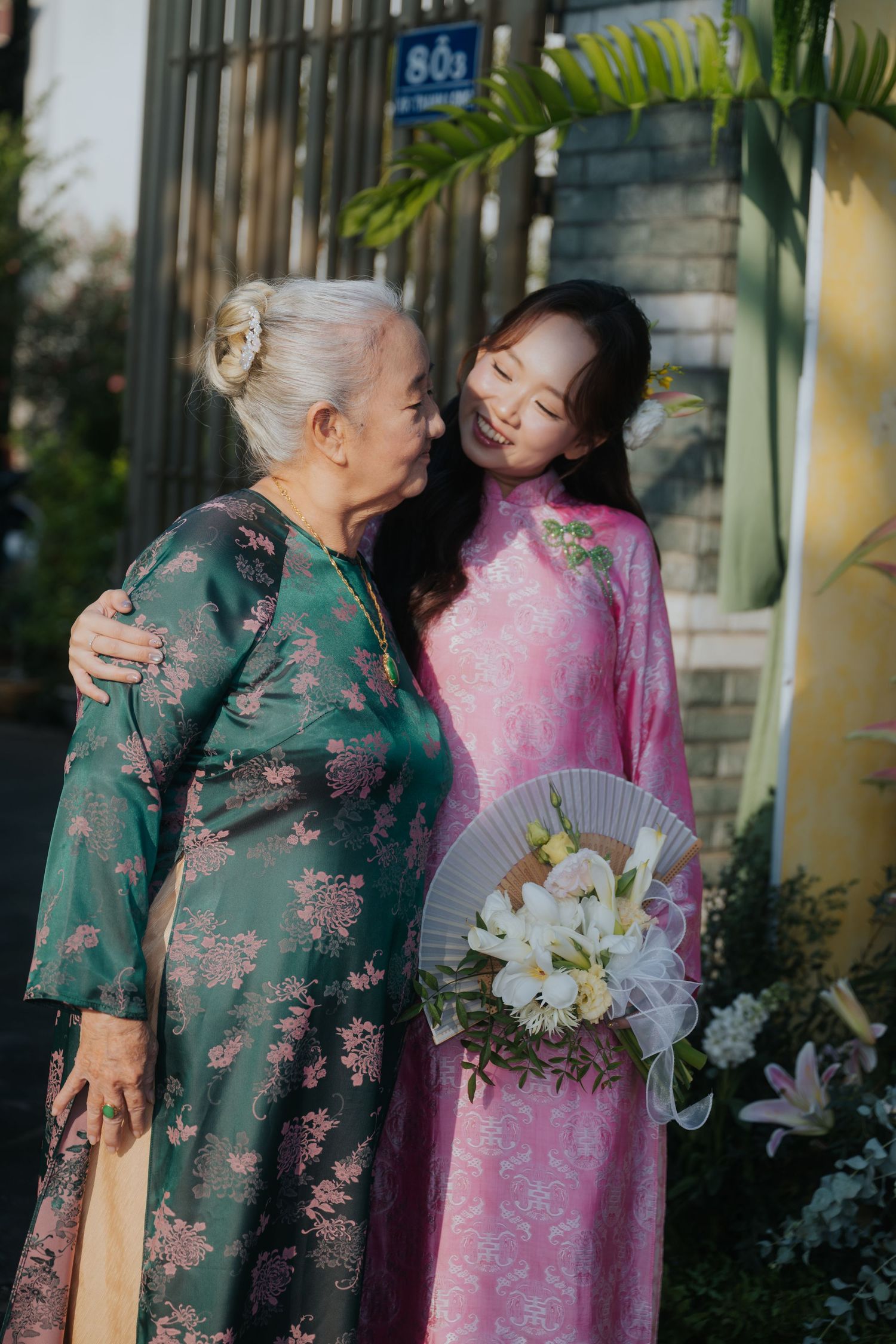 Two women in traditional Vietnamese ao dai dresses share a tender moment, one elderly in green, one young in pink holding flowers.