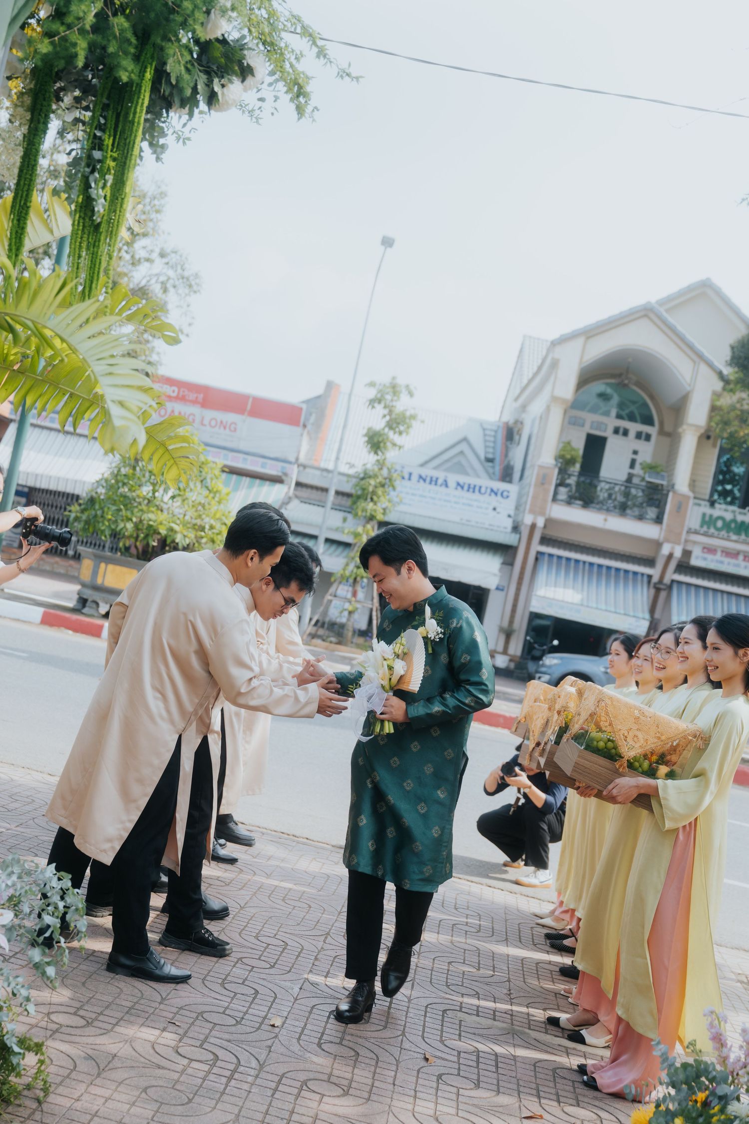 Vietnamese wedding ceremony with groom in green ao dai receiving guests outdoors on decorated street.