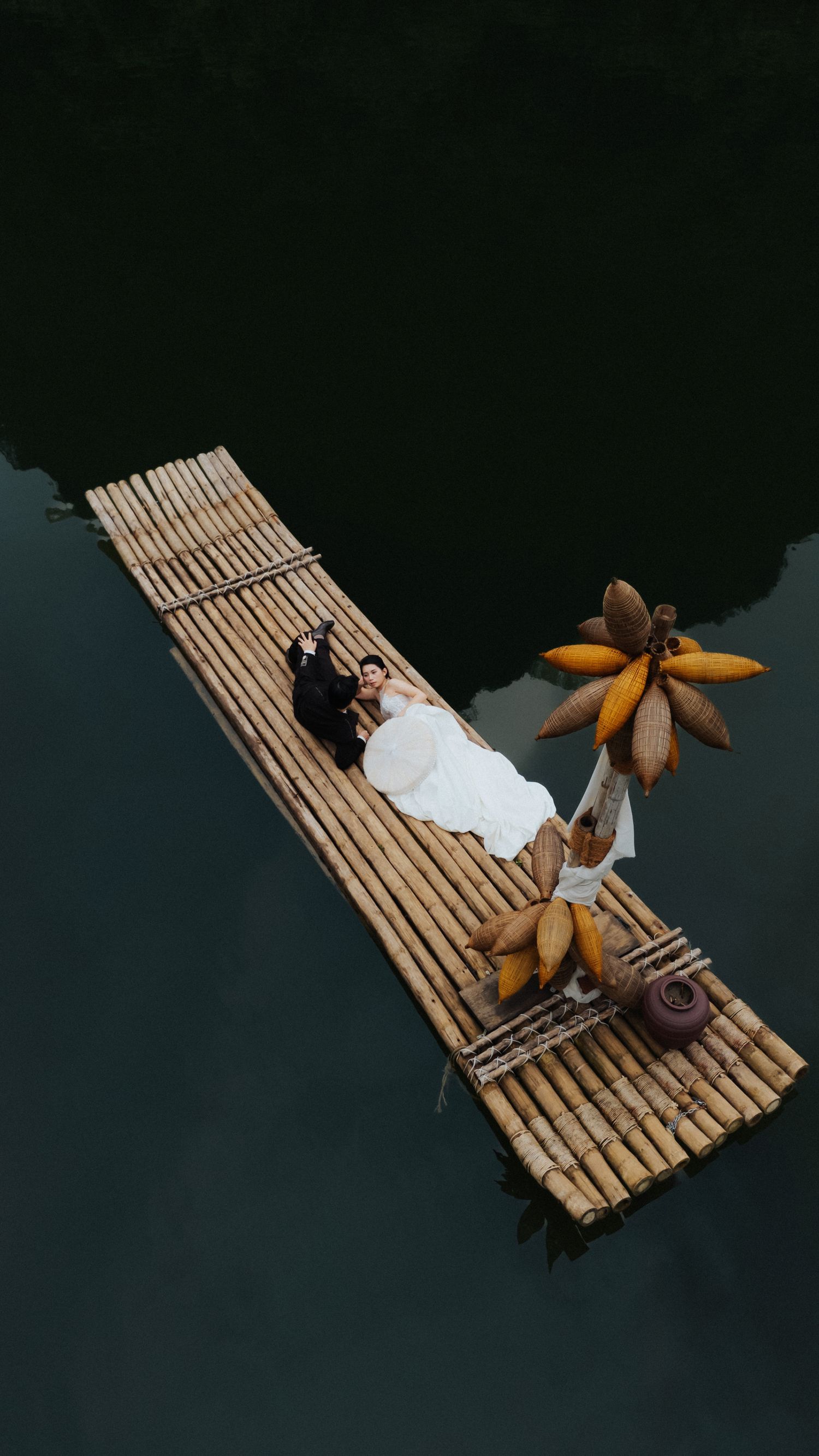 Aerial view of a couple in wedding attire lying on a bamboo raft floating on calm dark water with tropical fruit.