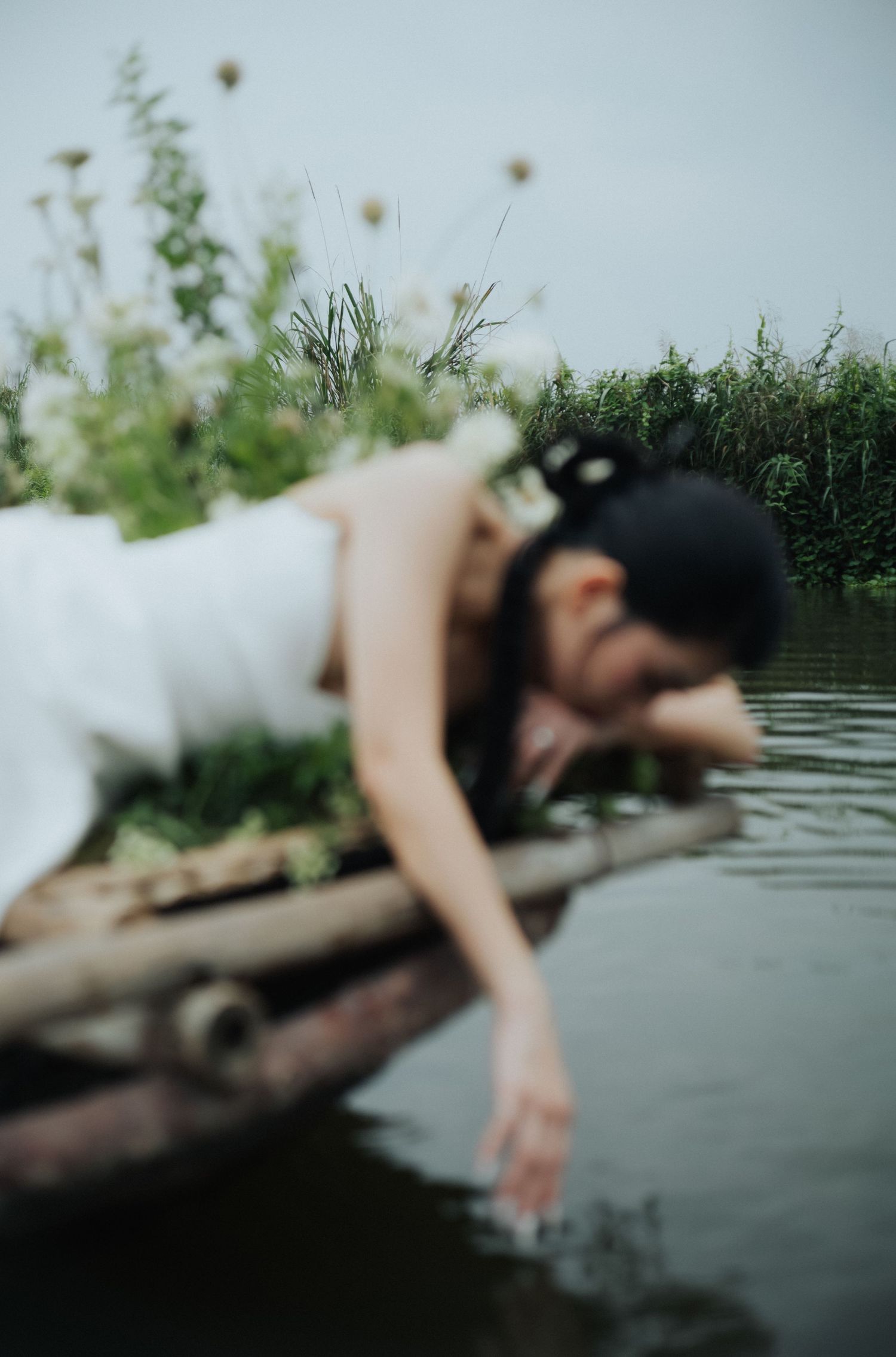 Woman in white dress lying on bamboo raft, reaching down to touch calm river water, surrounded by wildflowers.