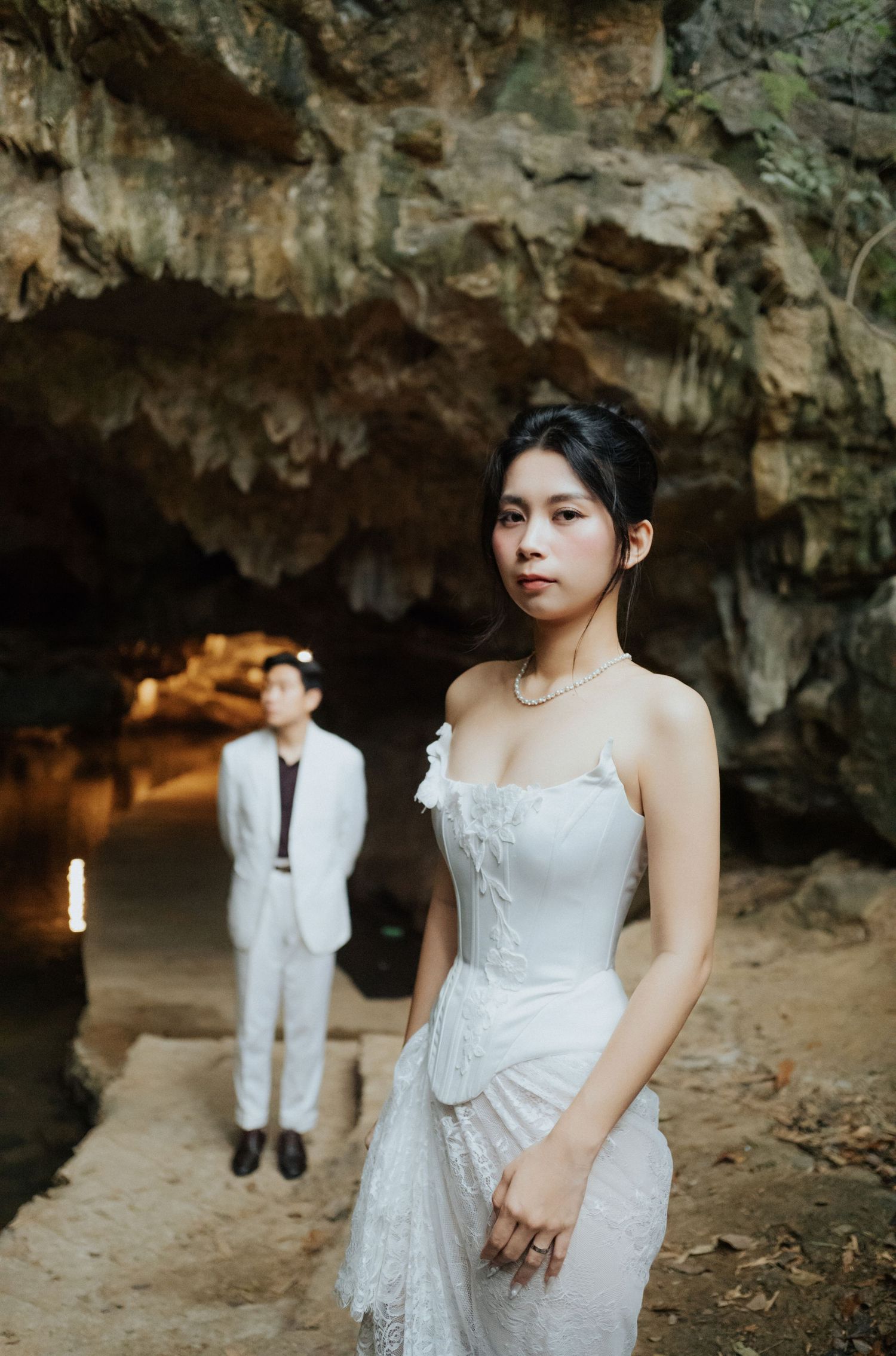 A bride in a white strapless gown poses in a cave with a groom in a white suit standing in the background.