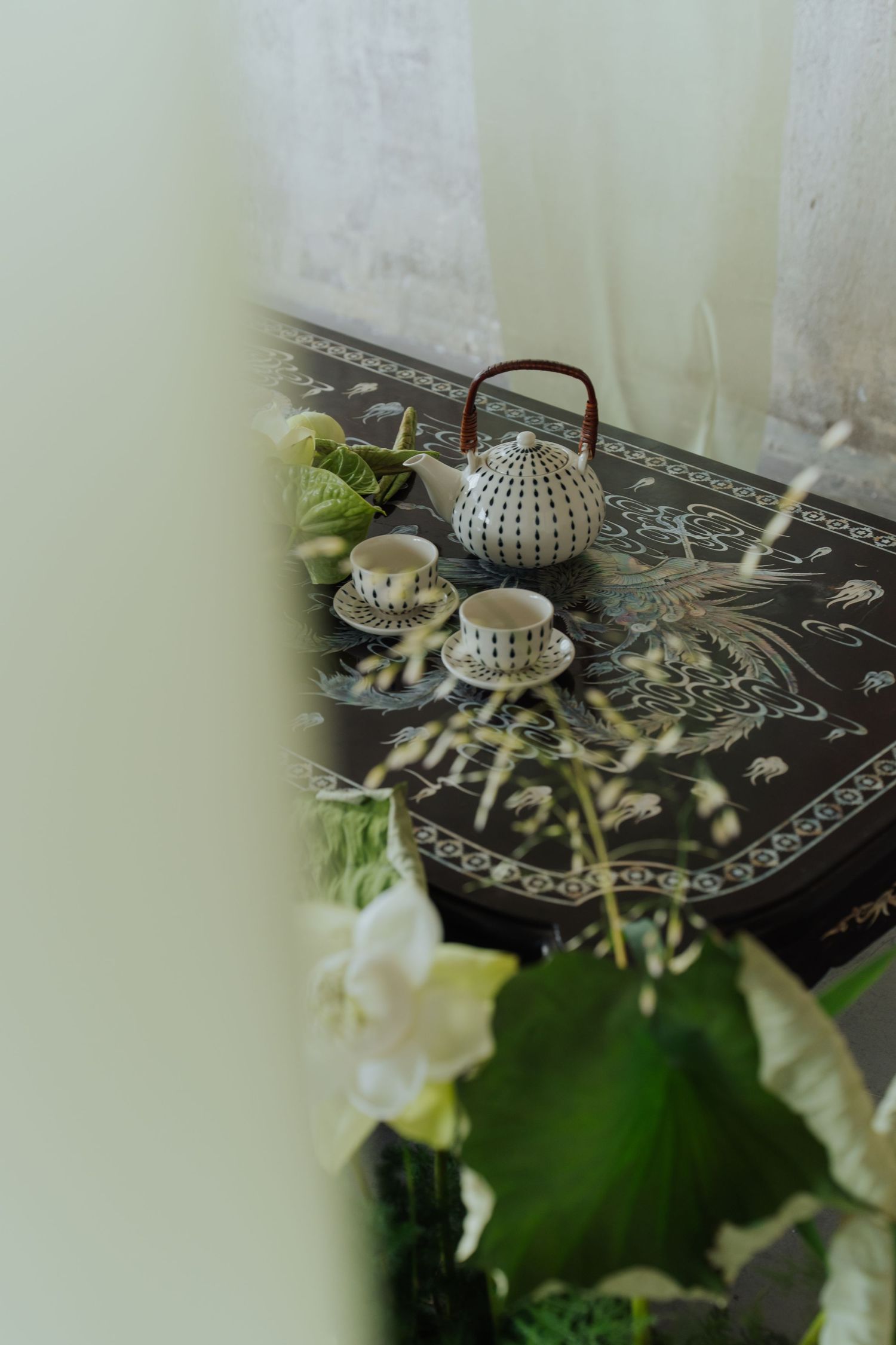 A decorative black Asian-style table with a white polka dot teapot and matching cups surrounded by lush green plants.