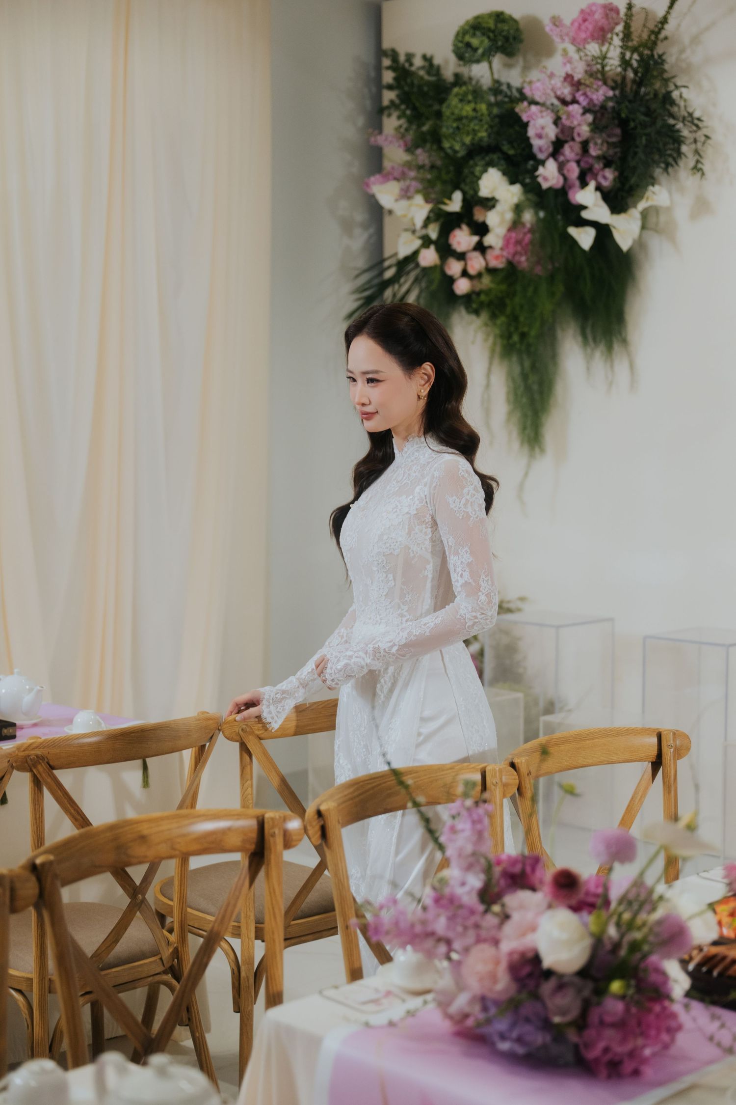 A woman in a white lace ao dai dress stands gracefully among wooden chairs at a floral-decorated wedding venue.