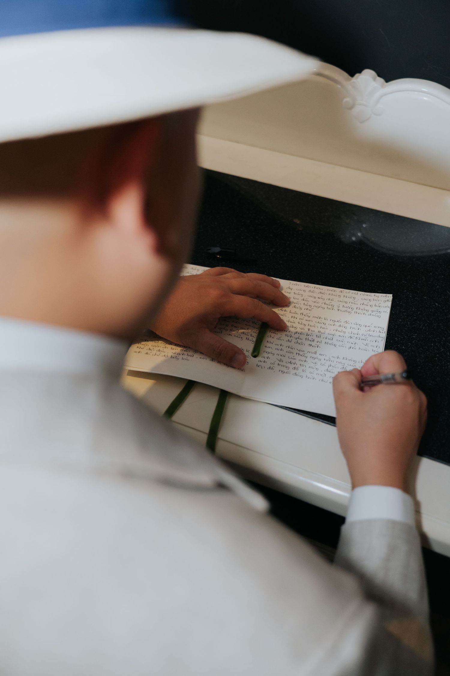 Person in white hat writing a handwritten letter on paper, viewed from above, wearing a ring and white shirt.