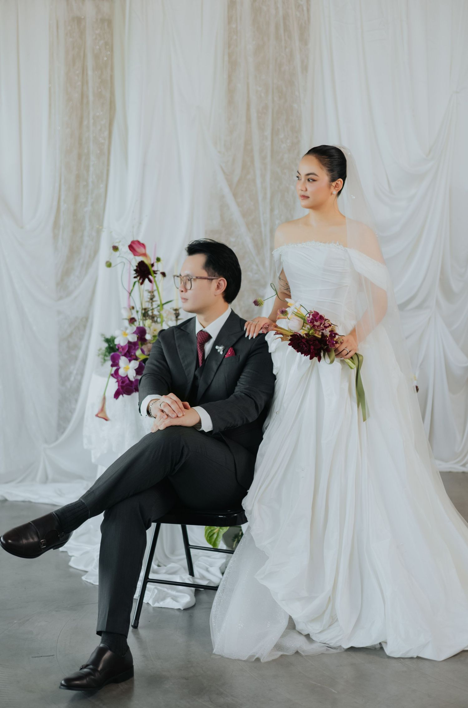 Elegant wedding couple posing in studio, groom seated in suit, bride standing in white off-shoulder gown holding burgundy bouquet.